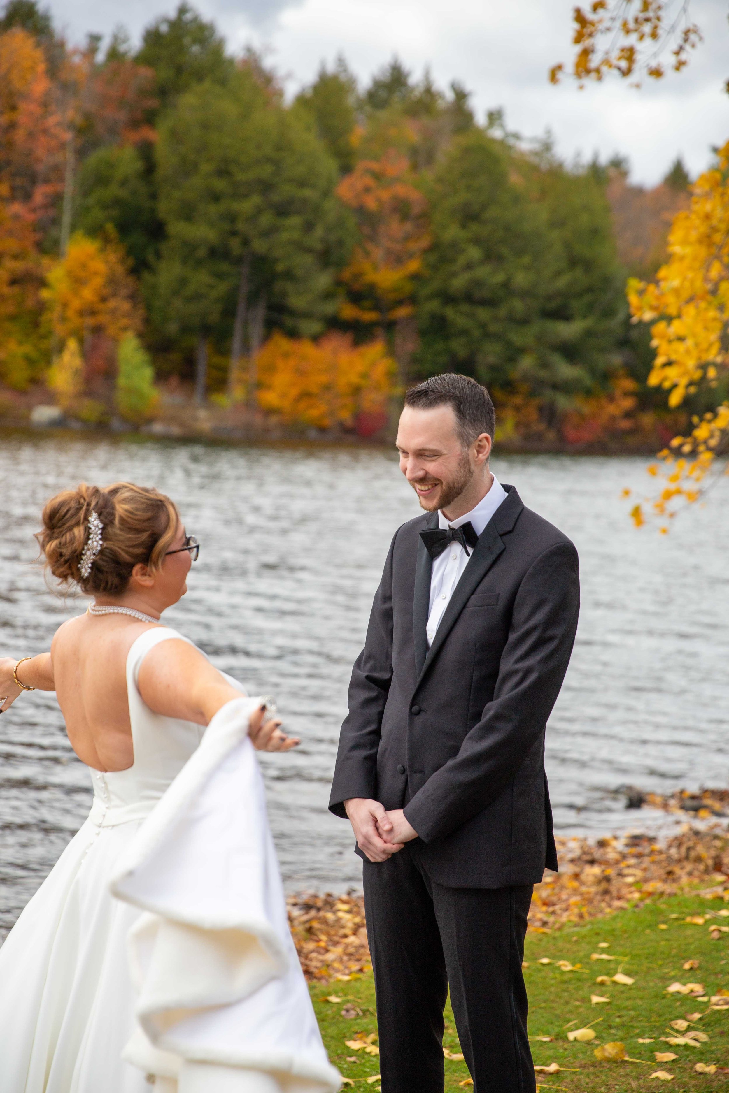 A bride and groom on their wedding day outdoors by a lake, with autumn trees and colorful fall foliage in the background.
