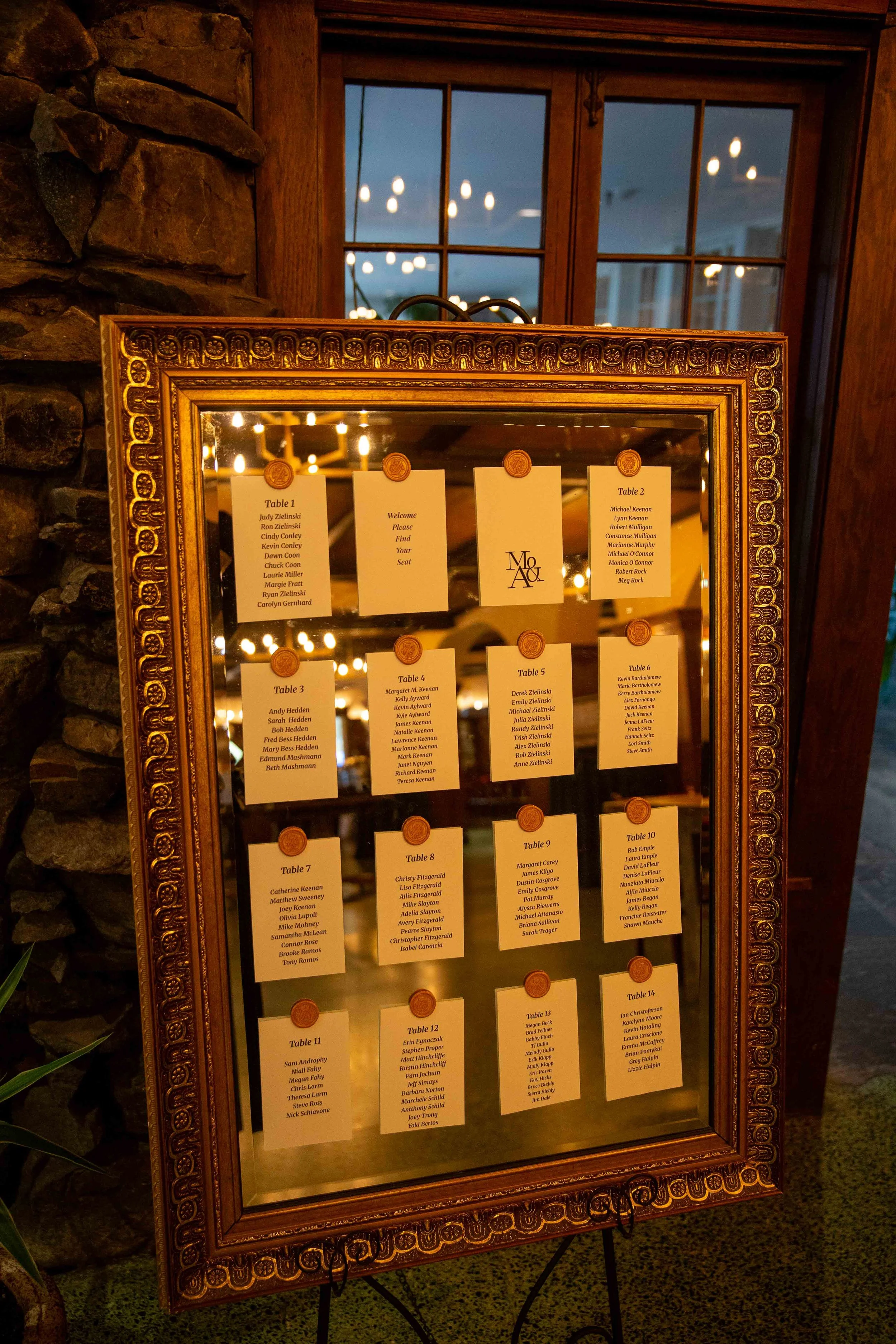 A framed seating chart with multiple guest names arranged by tables at an event. The frame is ornate, and the background shows a warmly lit room with a window and chandelier reflections.