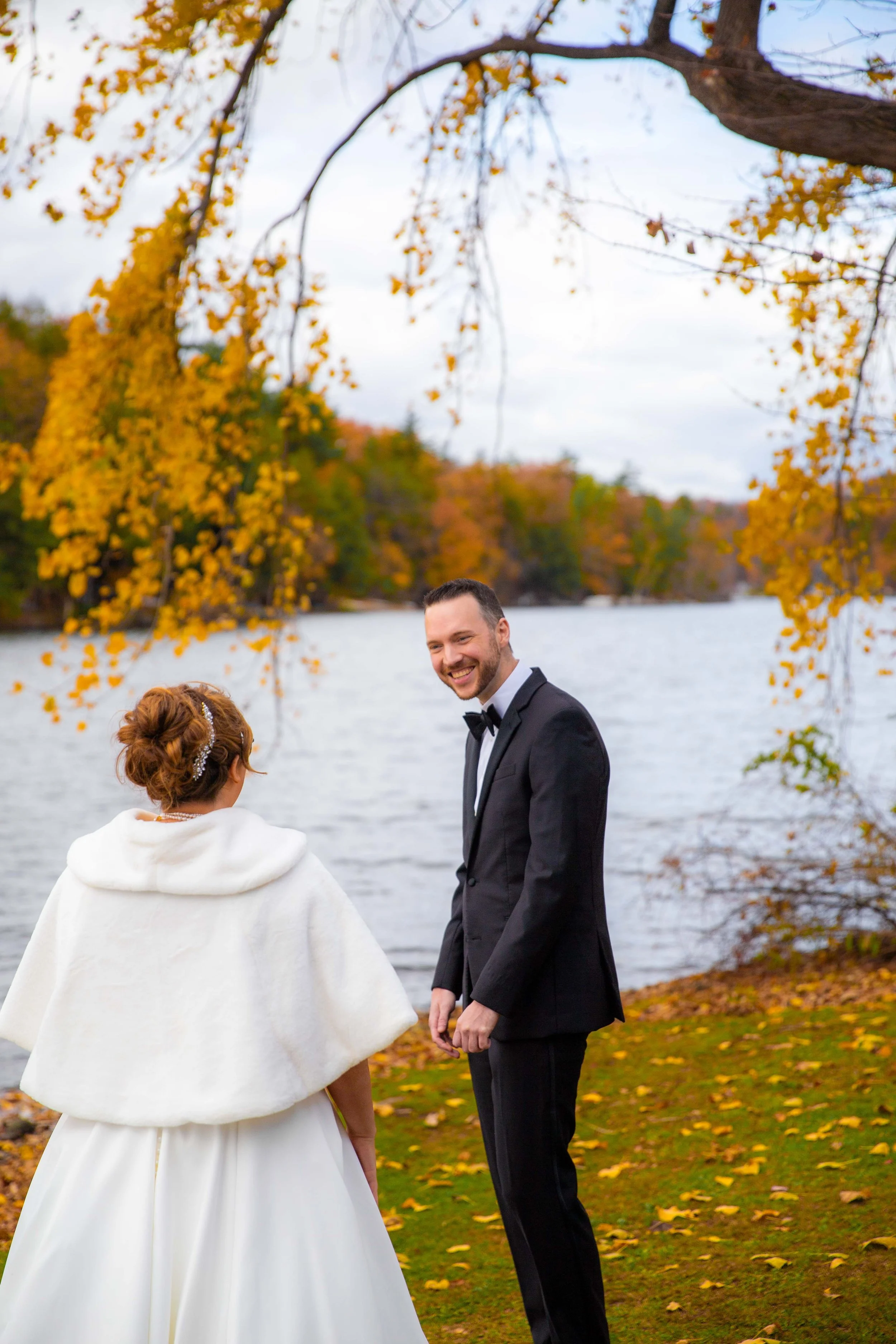 A bride and groom with wedding attire standing outdoors by a lake, surrounded by autumn trees with yellow and orange leaves.