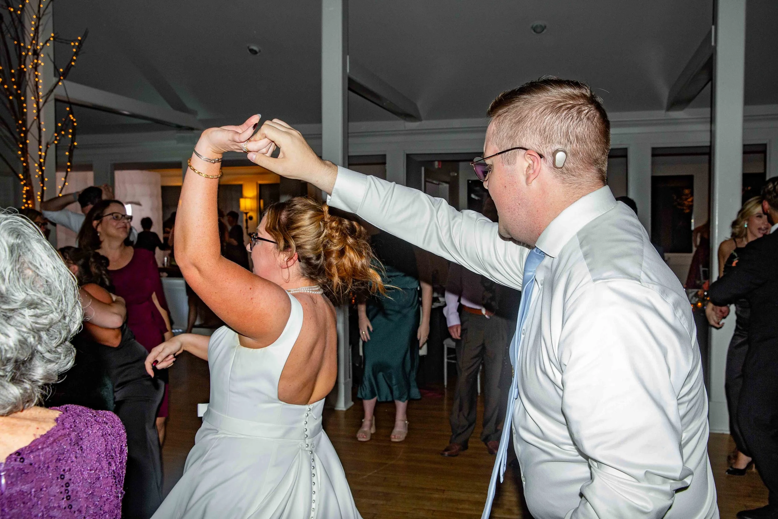 A couple dancing at a wedding reception. The woman wears a white dress with a low back, and the man wears a white shirt with a blue tie. They are holding hands as they dance. Other guests are visible in the background, enjoying the event.