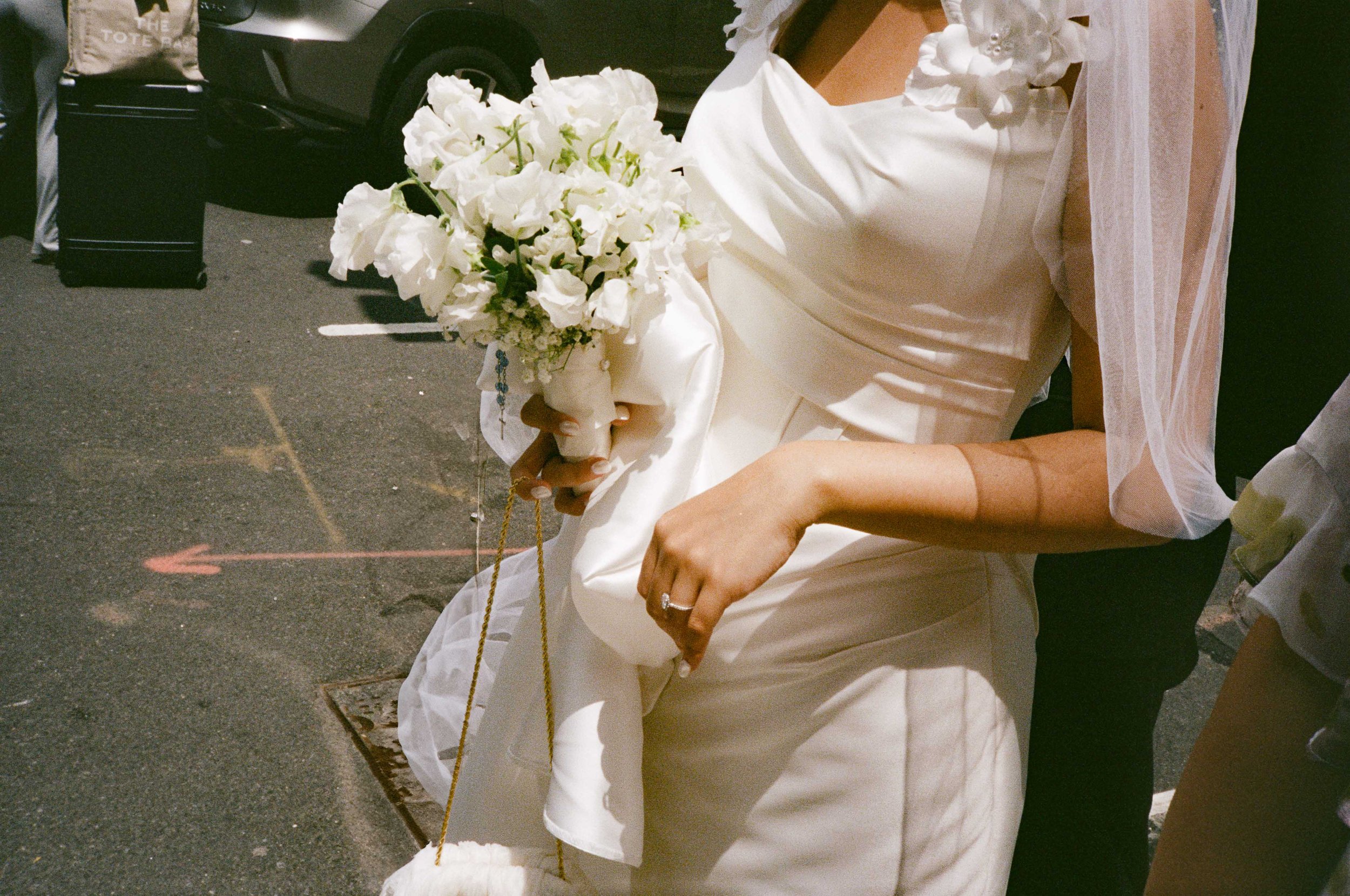 A woman dressed in a white wedding gown holding a bouquet of white flowers on a city street.