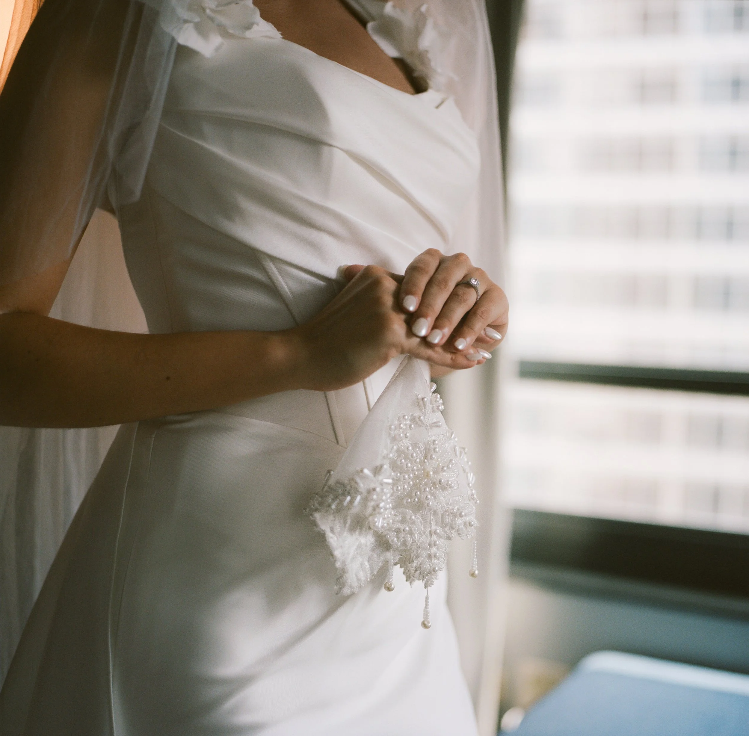 Close-up of a bride's hands with an engagement ring, holding a lace and beaded handkerchief, while wearing a white wedding dress near a window.