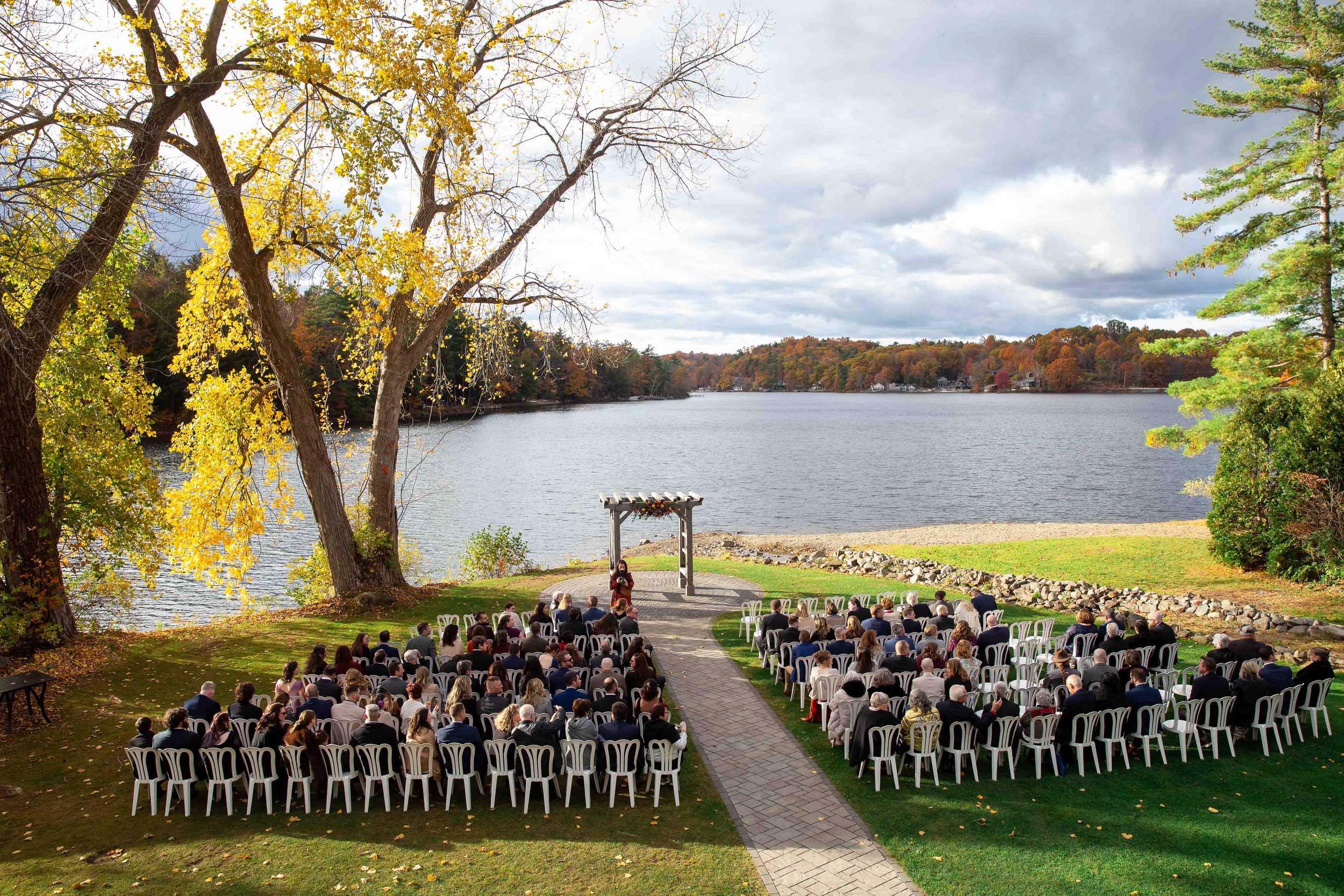 Outdoor wedding ceremony setup by a lake in autumn, with guests seated on white chairs, an arch decorated with flowers, and colorful trees in the background.