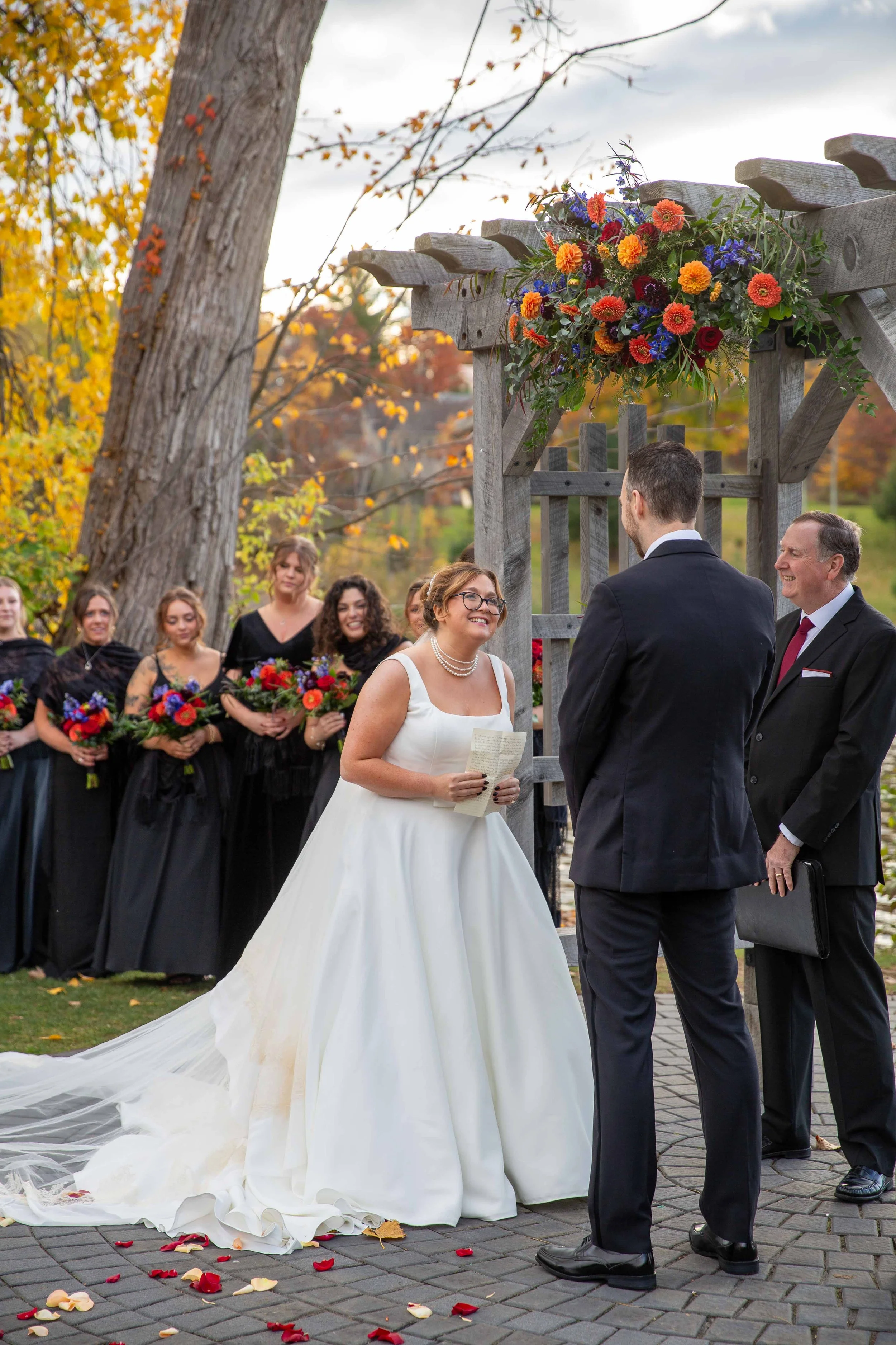A wedding ceremony outdoors with a bride in a white gown and pearls, a groom in a dark suit, and a priest. Bridesmaids in black dresses holding bouquets, standing in the background among autumn trees. Colorful flowers decorate a wooden archway.