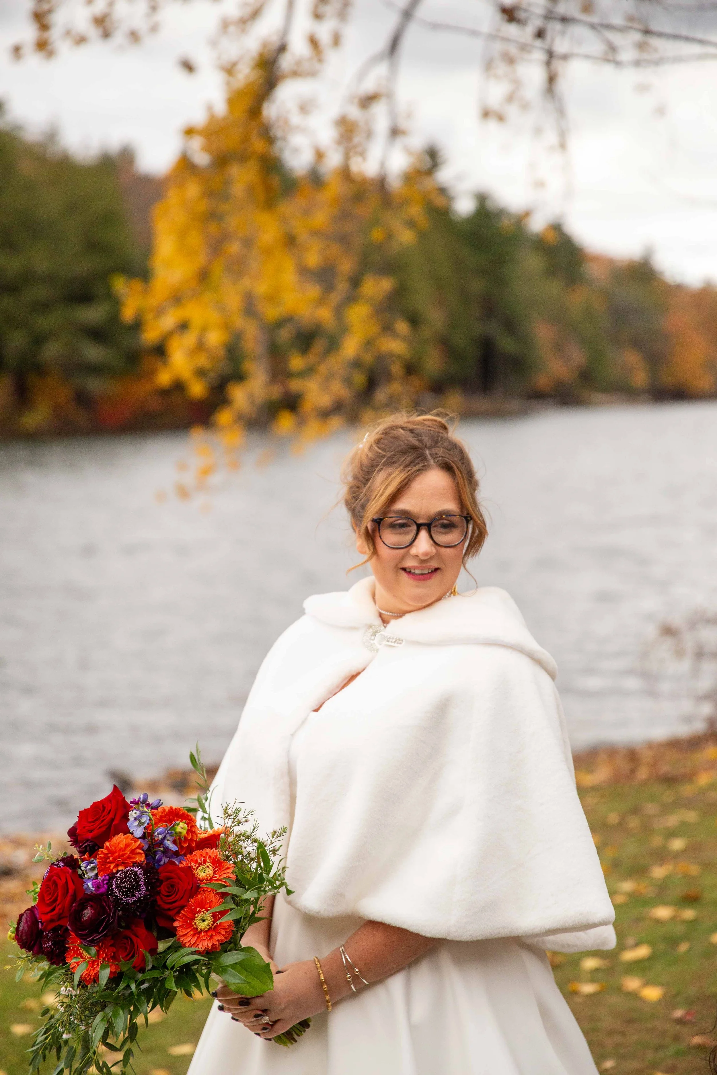 A smiling woman in a white dress and cape holding a bouquet of red, purple, and orange flowers by a lake during autumn, with colorful fall foliage in the background.