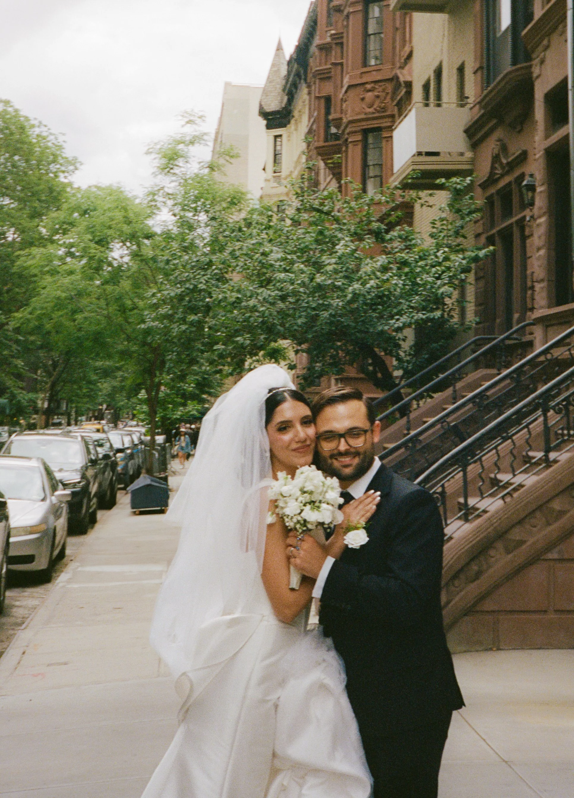 A bride and groom standing close together on a city sidewalk, smiling at the camera. The bride wears a white wedding dress and veil, holding a bouquet of white flowers. The groom is dressed in a black suit with glasses. Behind them are brownstone bui
