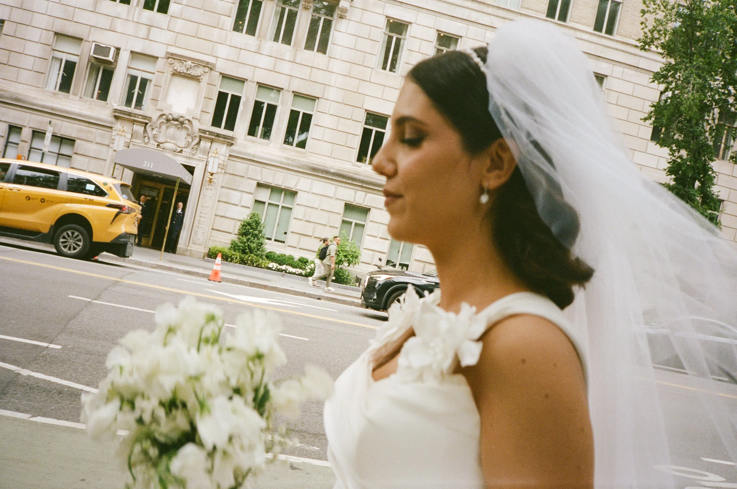 A bride standing on a city sidewalk holding a bouquet of white flowers, wearing a white wedding dress and veil, with a city building and yellow taxi in the background.