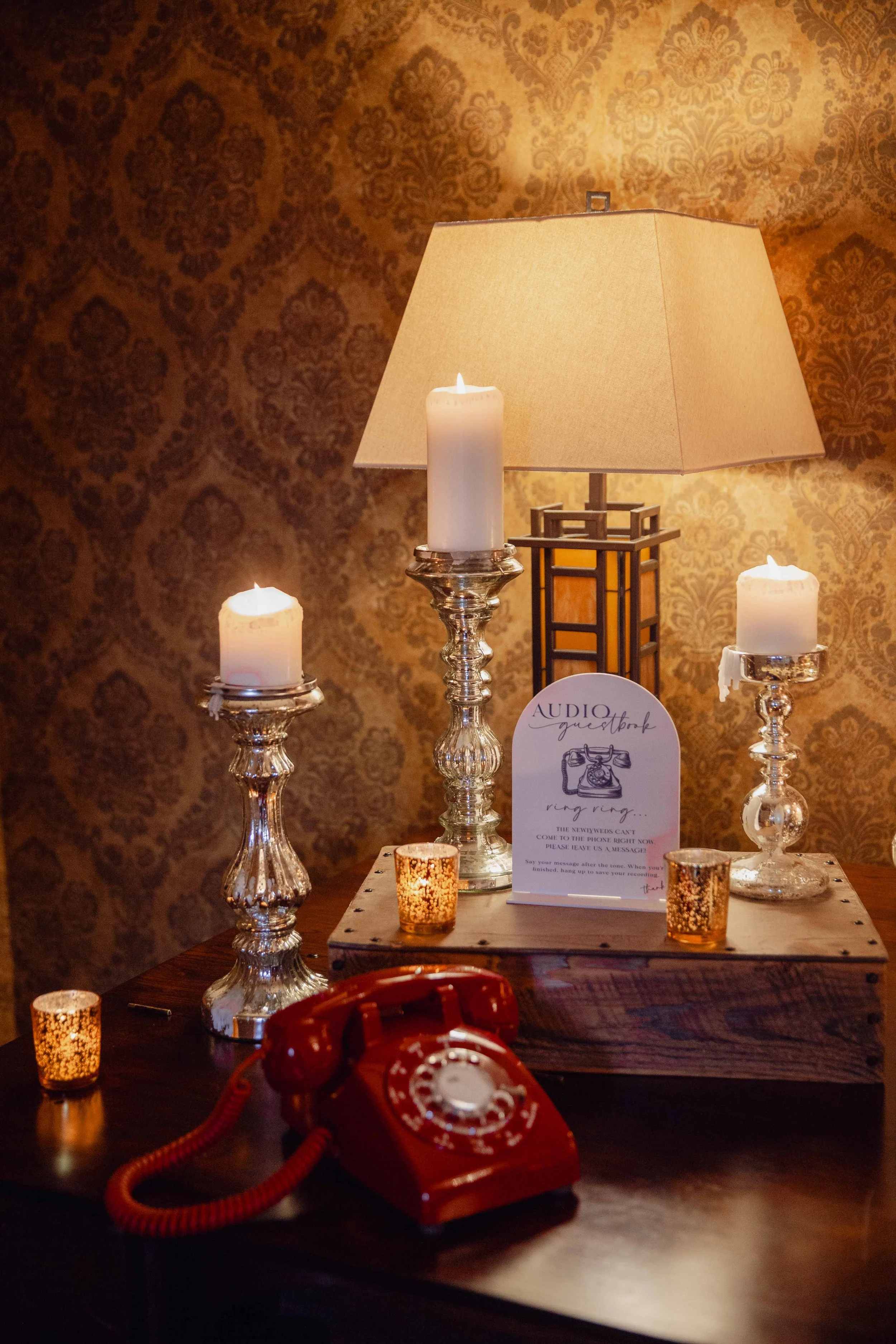 Decorative table setup with candles, a lamp, and a vintage red rotary telephone against a patterned wallpaper background.