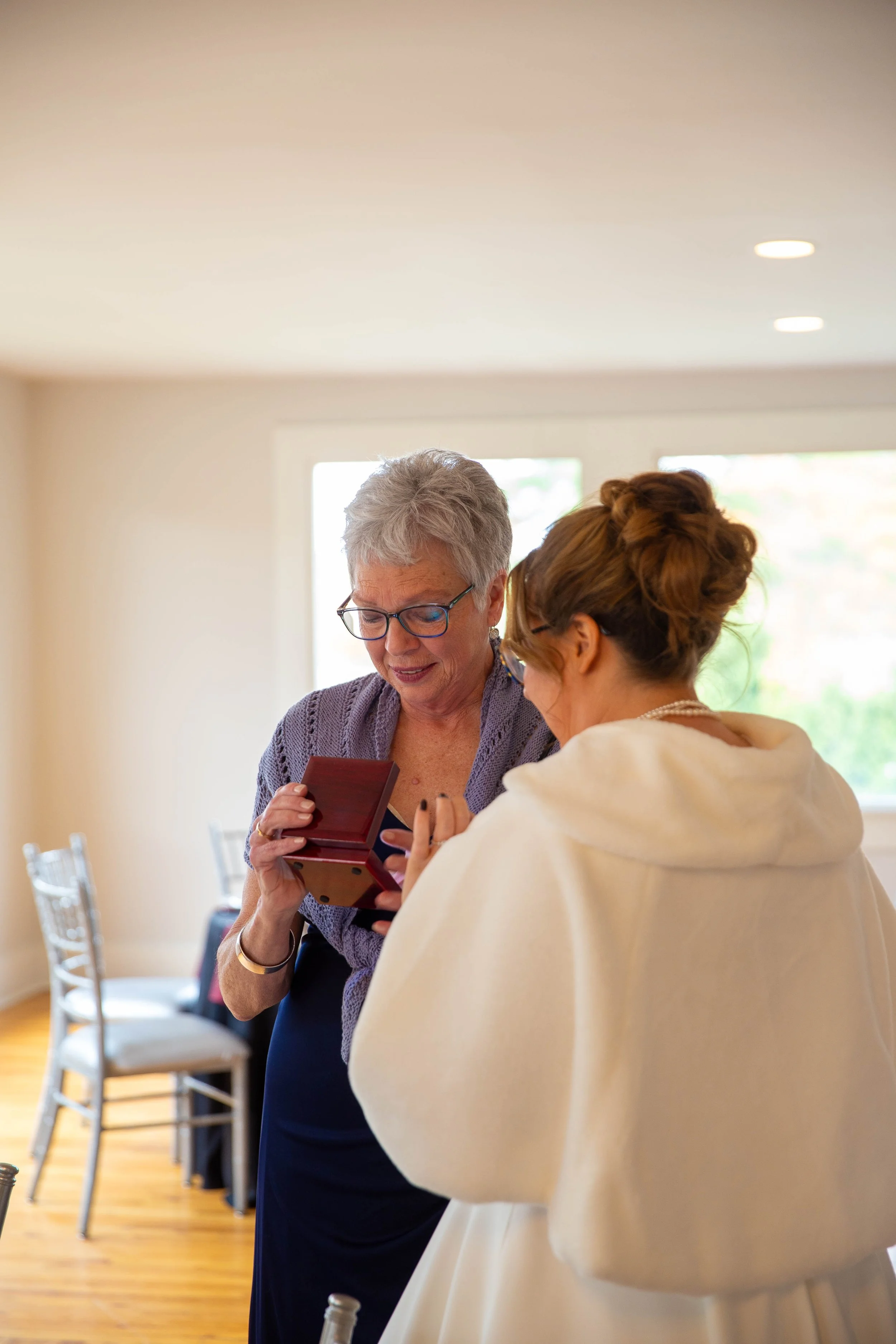 An elderly woman with glasses and gray hair is holding a small open box and smiling as she talks to a younger woman with a bun hairstyle and a white shawl.