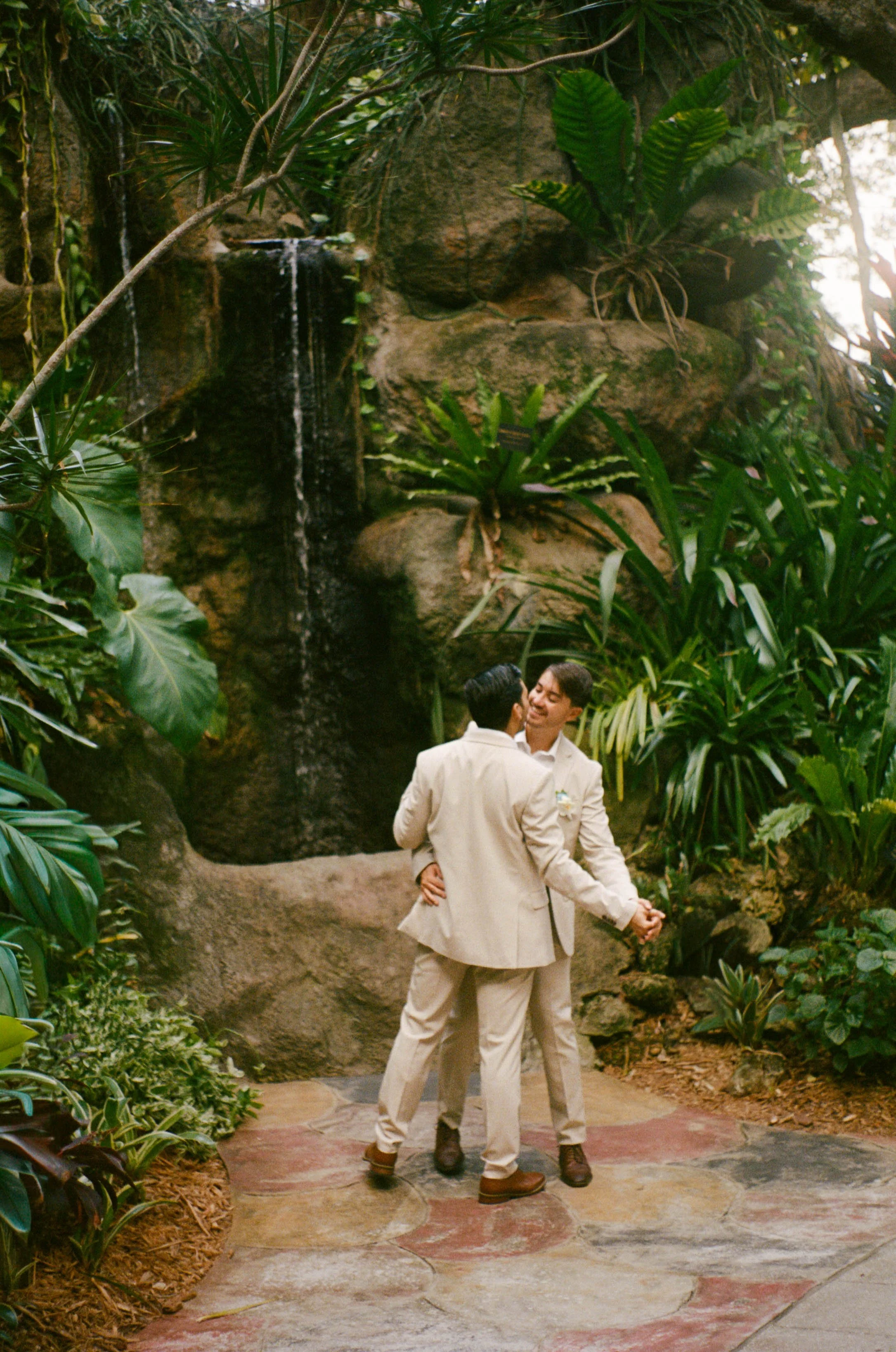 Two men in beige suits are dancing in a lush indoor garden with a small waterfall and large green plants.