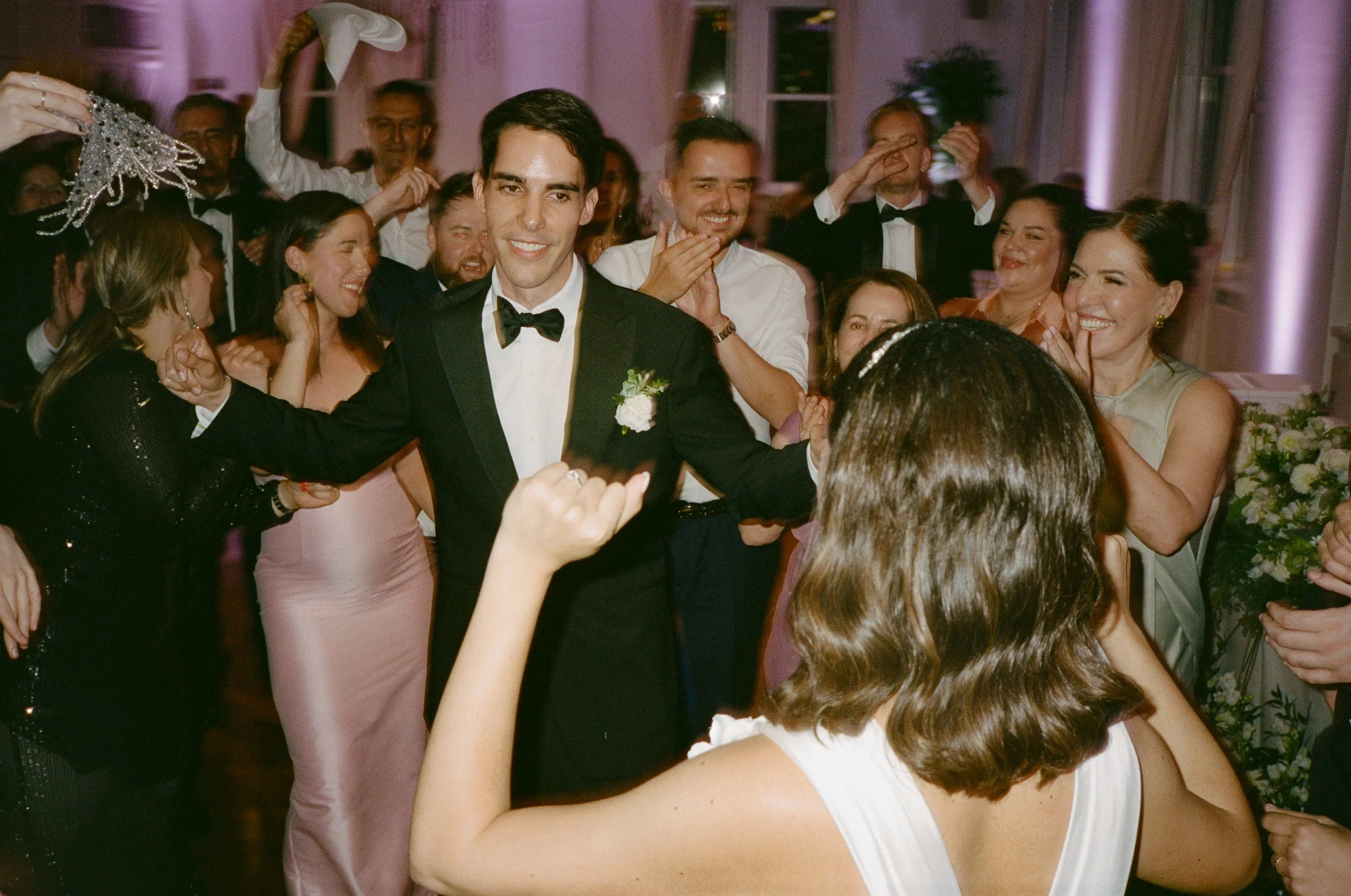 A groom in a tuxedo with a bow tie and boutonniere, smiling and celebrating with family and friends at a wedding reception. The scene is lively, with guests dancing and enjoying the moment.