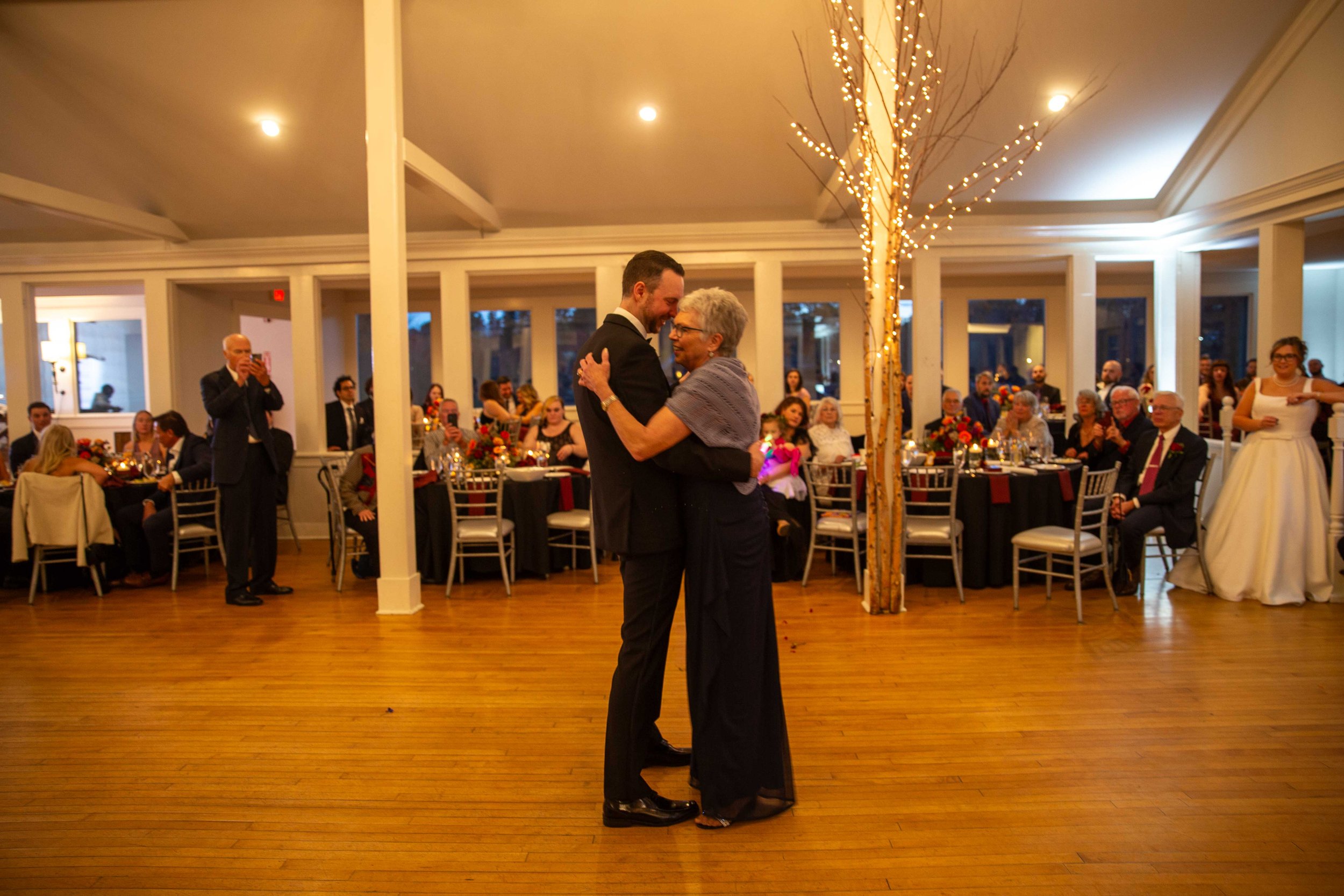 A young man and an older woman dancing together at a formal event in a decorated ballroom with guests seated at tables in the background.