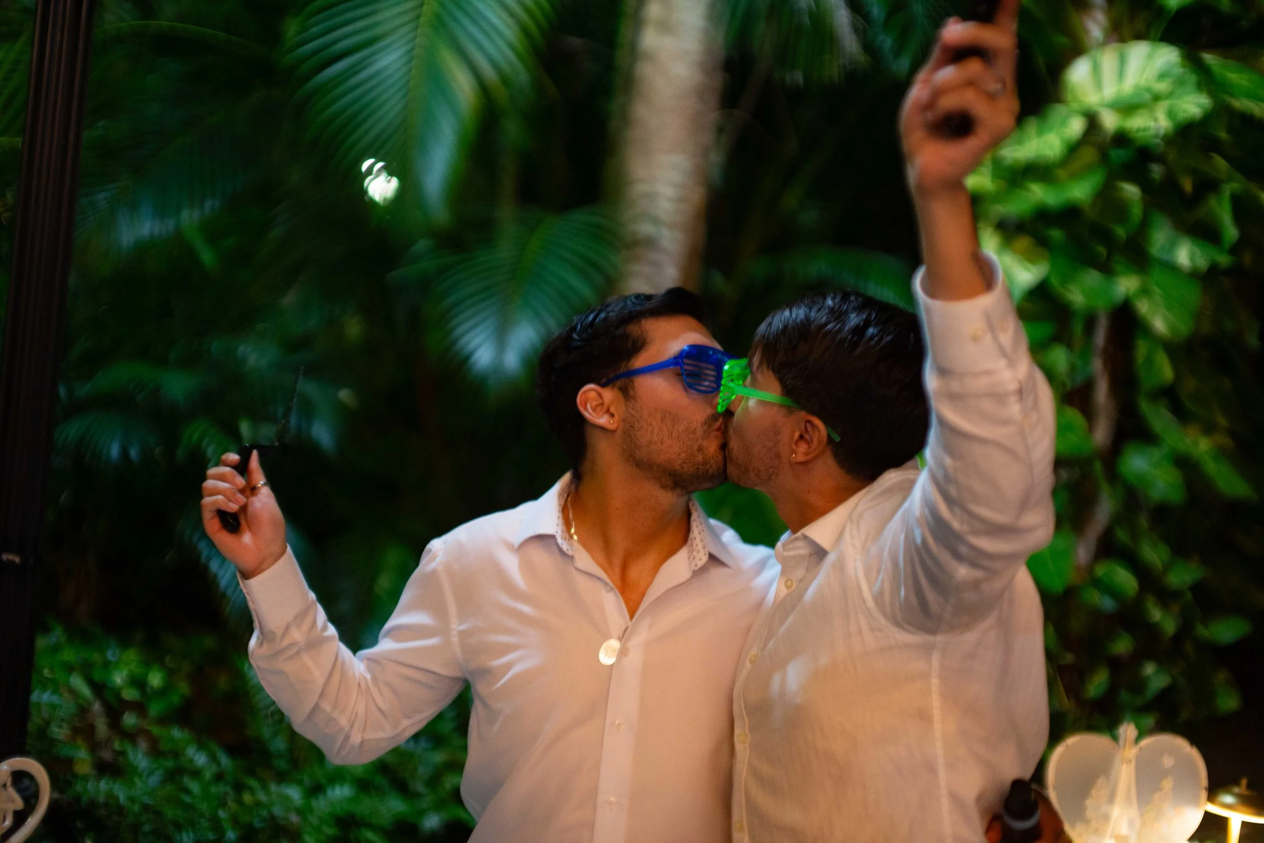 Two men in white shirts wearing colorful glasses kiss each other in a lush, green indoor setting.