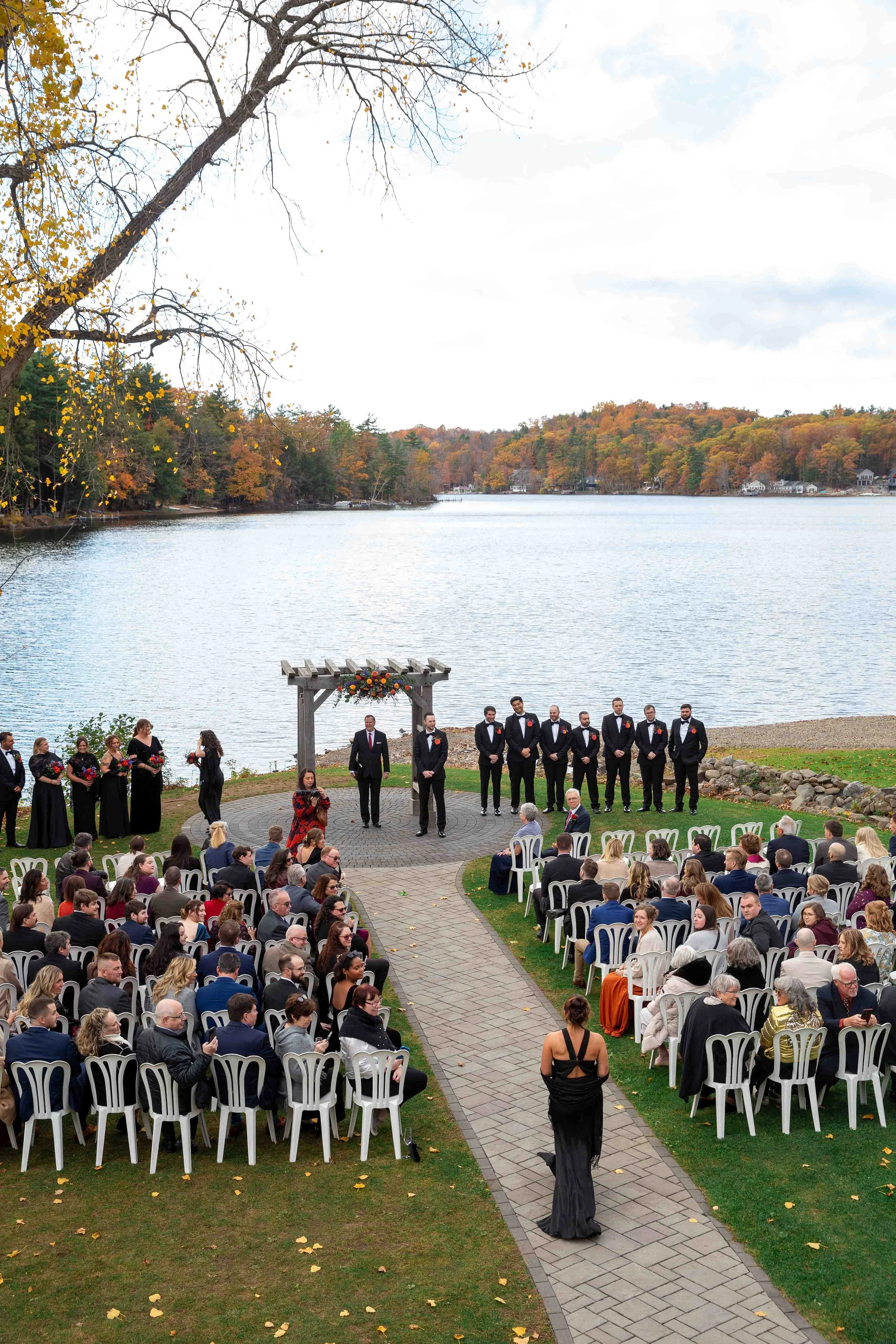 A wedding ceremony taking place outdoors by a lake, with guests seated and a bridal party standing under a wooden arch decorated with flowers.