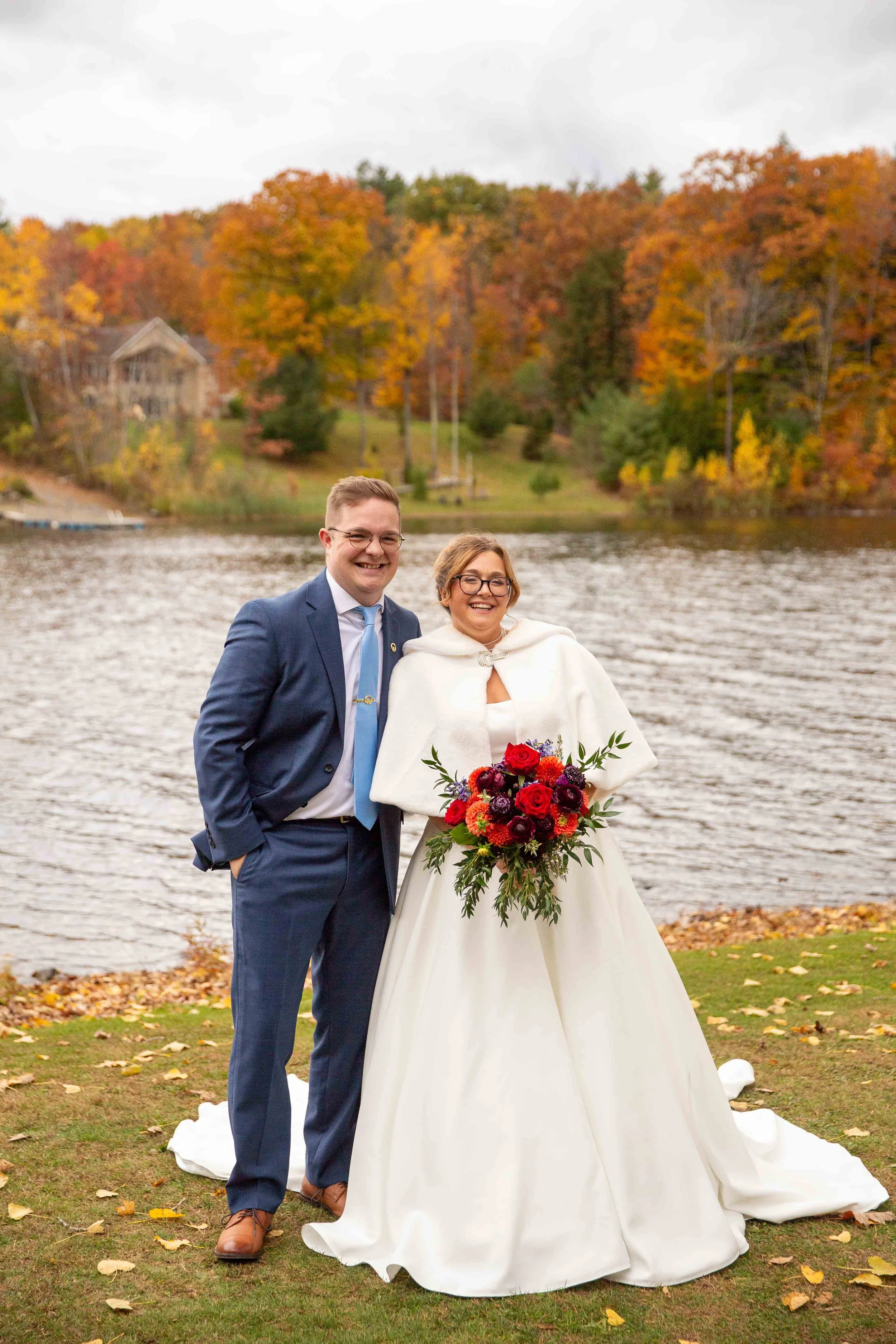 A bride and groom smiling outdoors near a lake with autumn trees in the background. The bride is wearing a white gown and holding a bouquet of red, purple, and orange flowers; the groom is in a blue suit and tie.