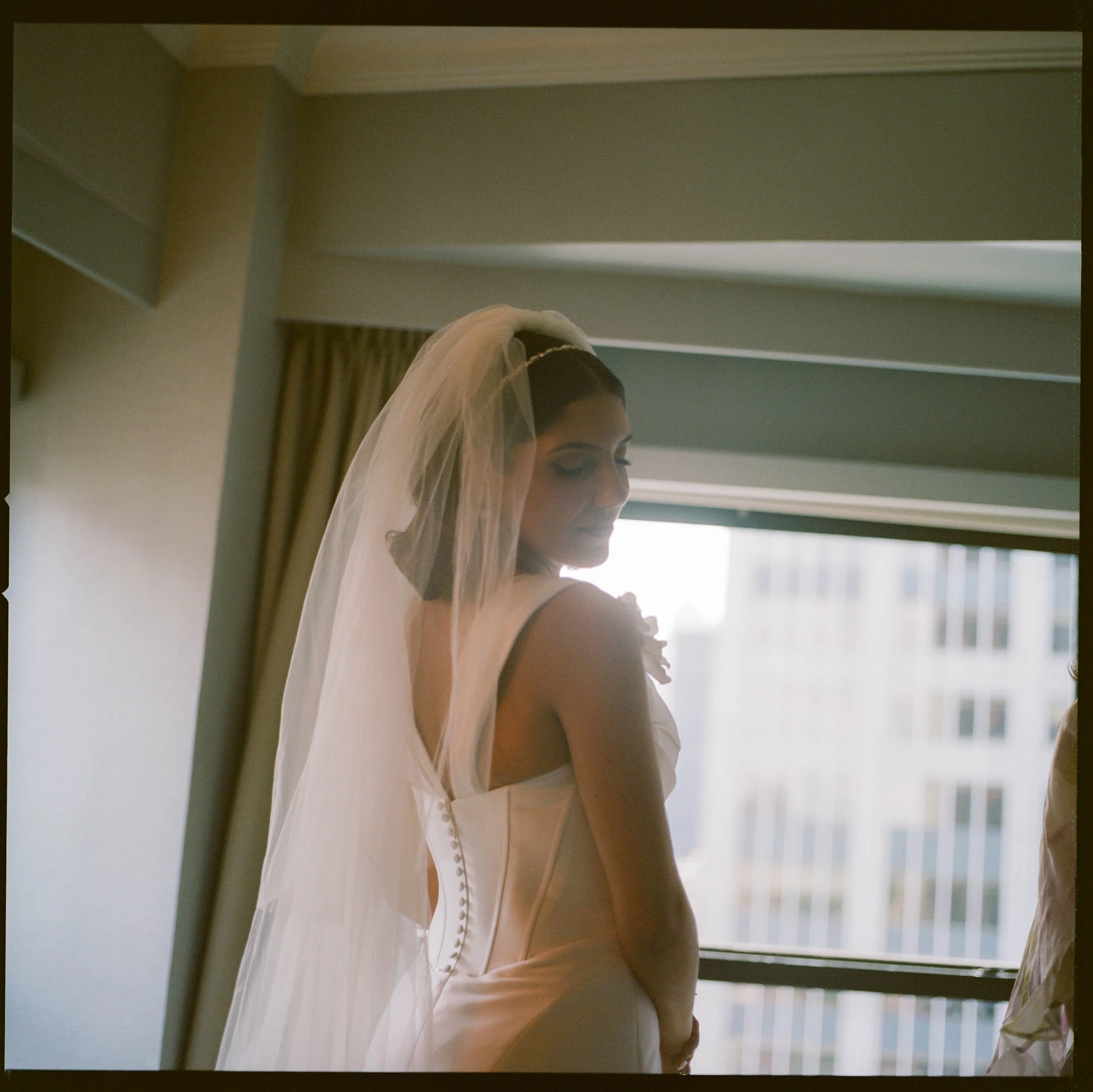 A bride wearing a white wedding dress and veil stands by a window with a cityscape view.