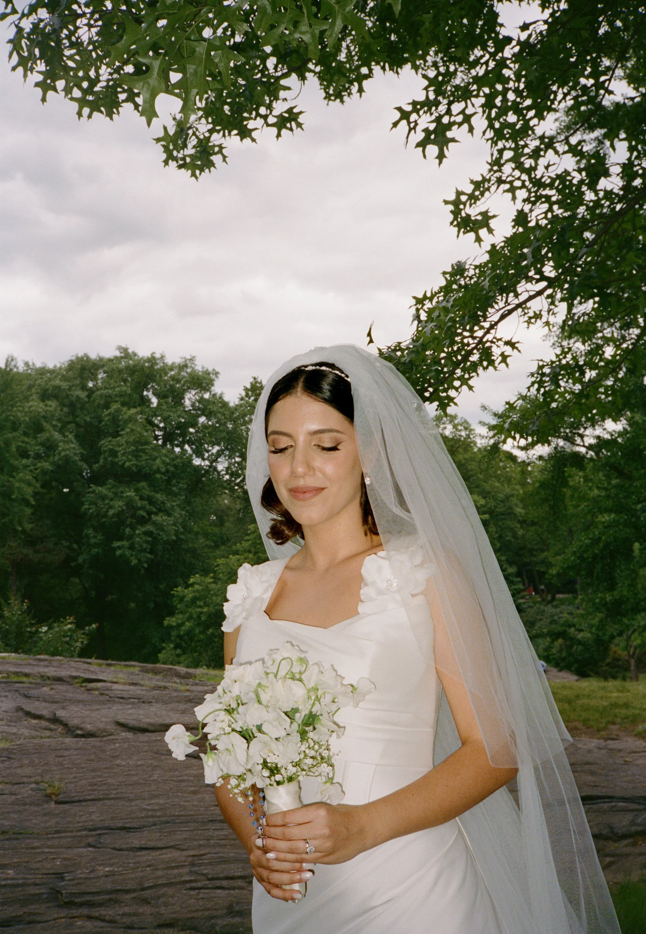 A bride with a veil and white dress holding a bouquet of white flowers in an outdoor setting with trees and cloudy sky.