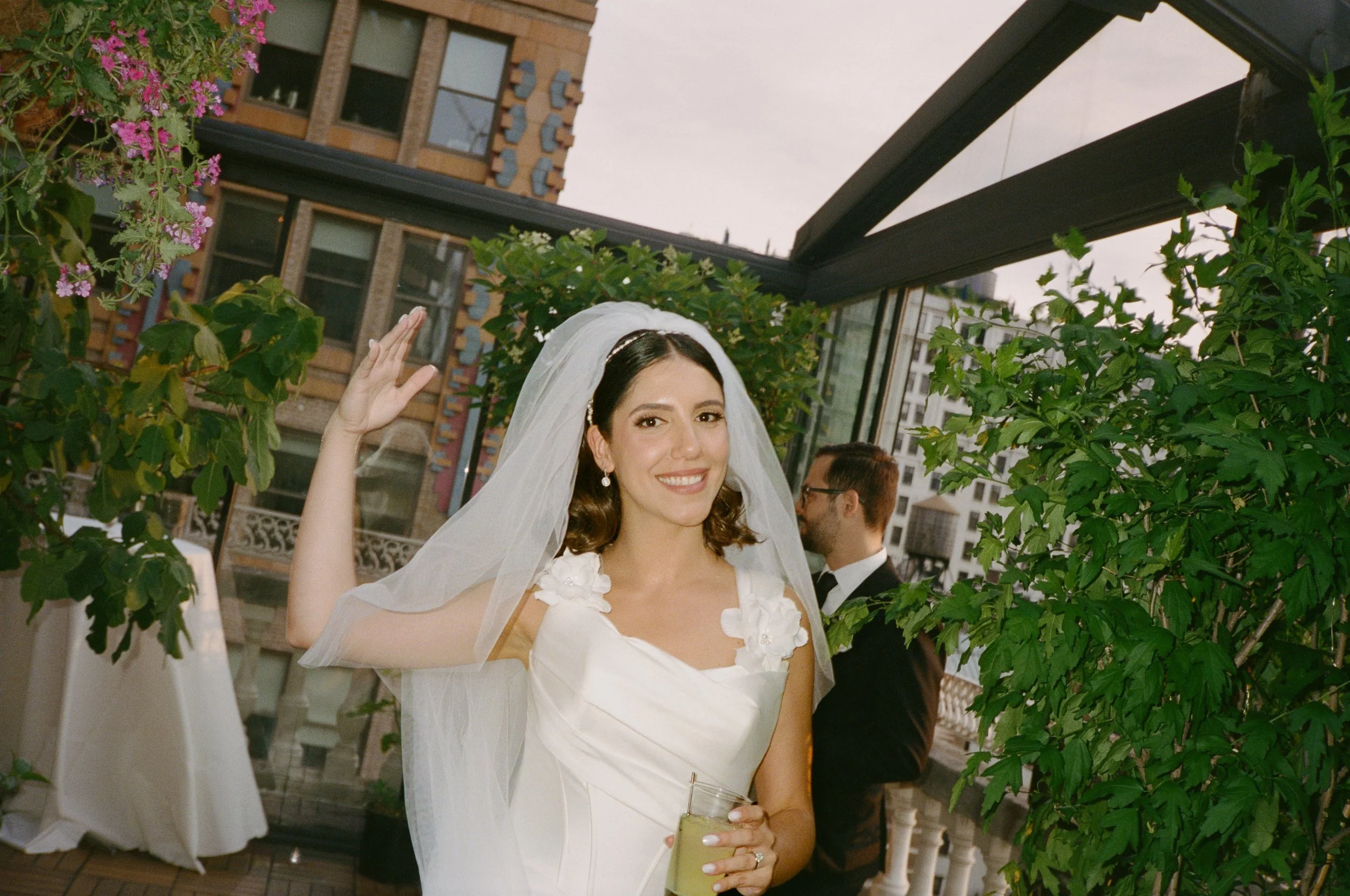 A bride with a white dress and veil smiling and waving at a wedding reception, holding a drink, with greenery and a cityscape background.