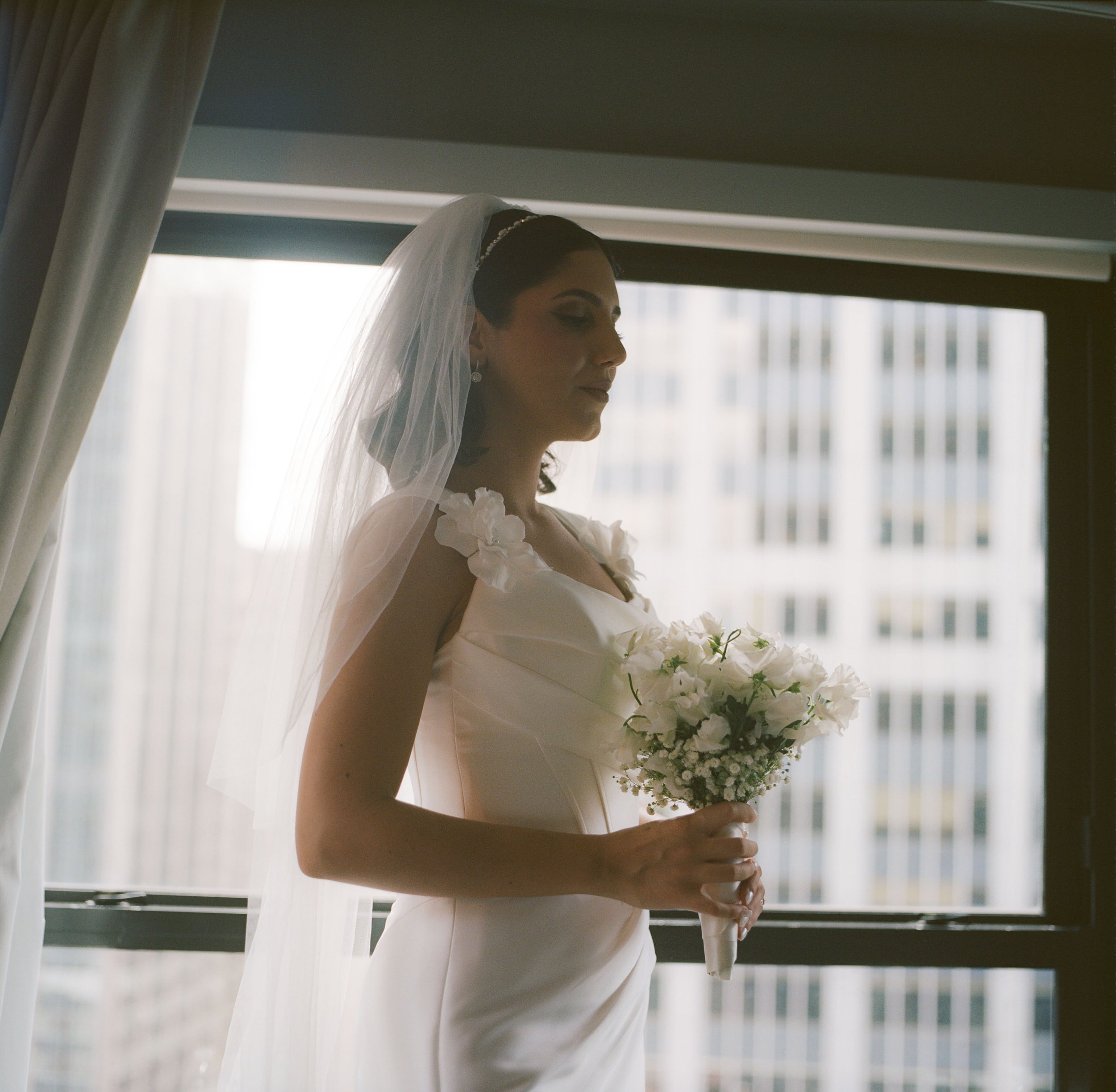Bride in a white wedding dress with floral accents on the shoulders, holding a bouquet of white flowers, standing inside near a window with cityscape background, wearing a veil and earrings.