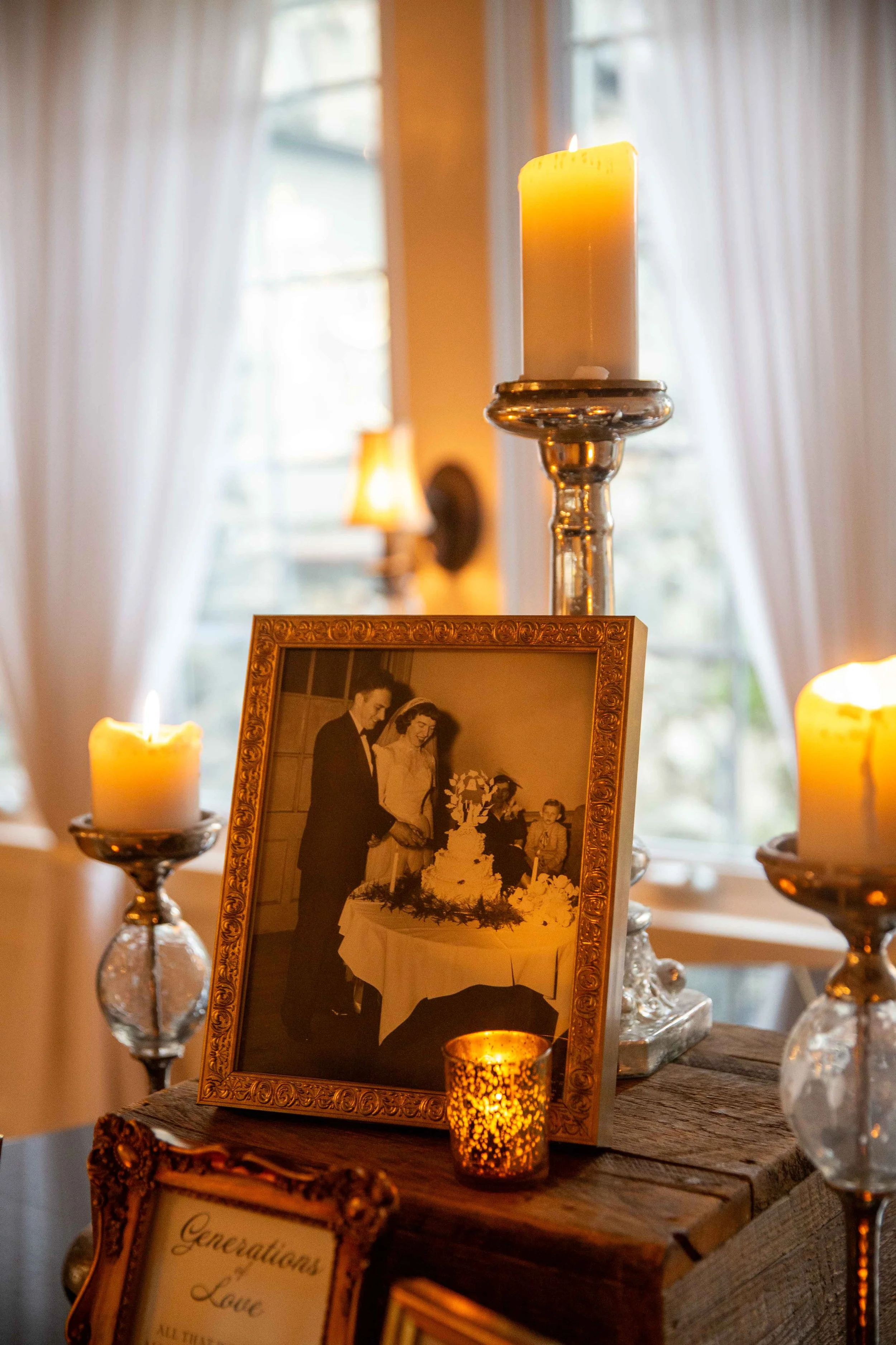 A framed wedding photo on a rustic wooden table, surrounded by lit candles in glass holders, with curtains and windows in the background.
