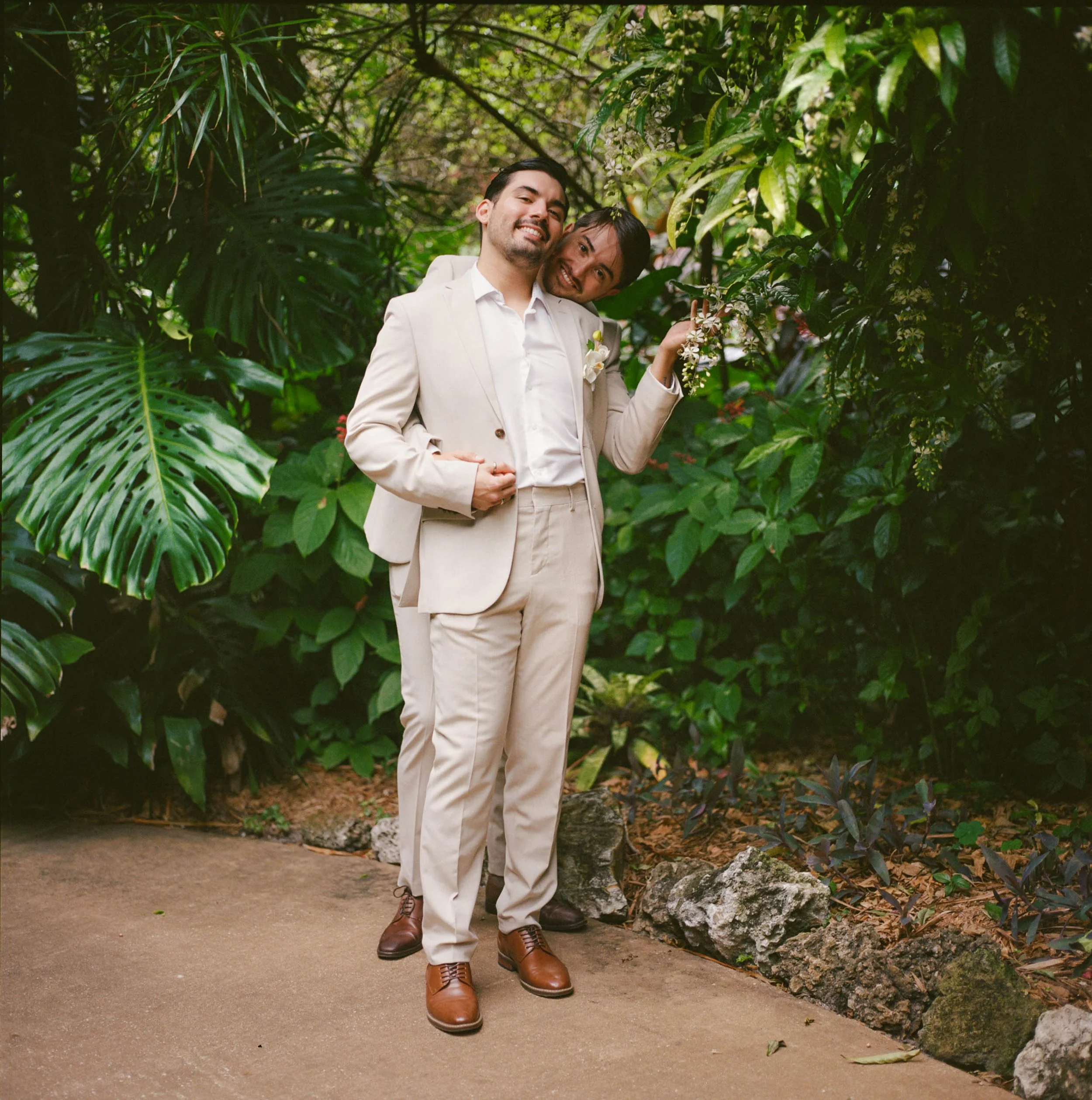 Two men dressed in beige suits standing in lush green garden, smiling and embracing, with one holding a small branch with white flowers.
