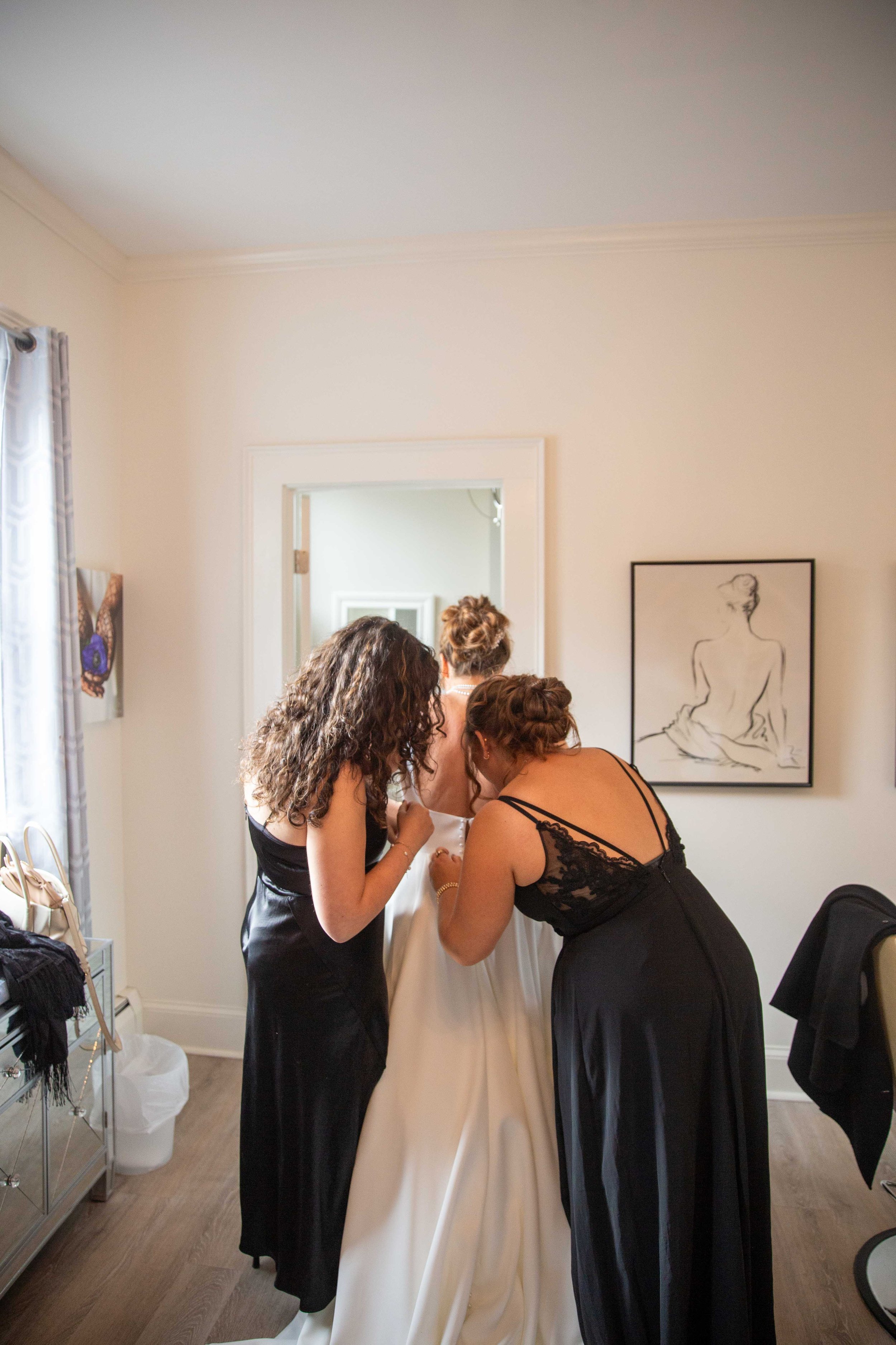 Three women, one bride in a wedding dress, getting ready in a bright room with wall art and natural light.