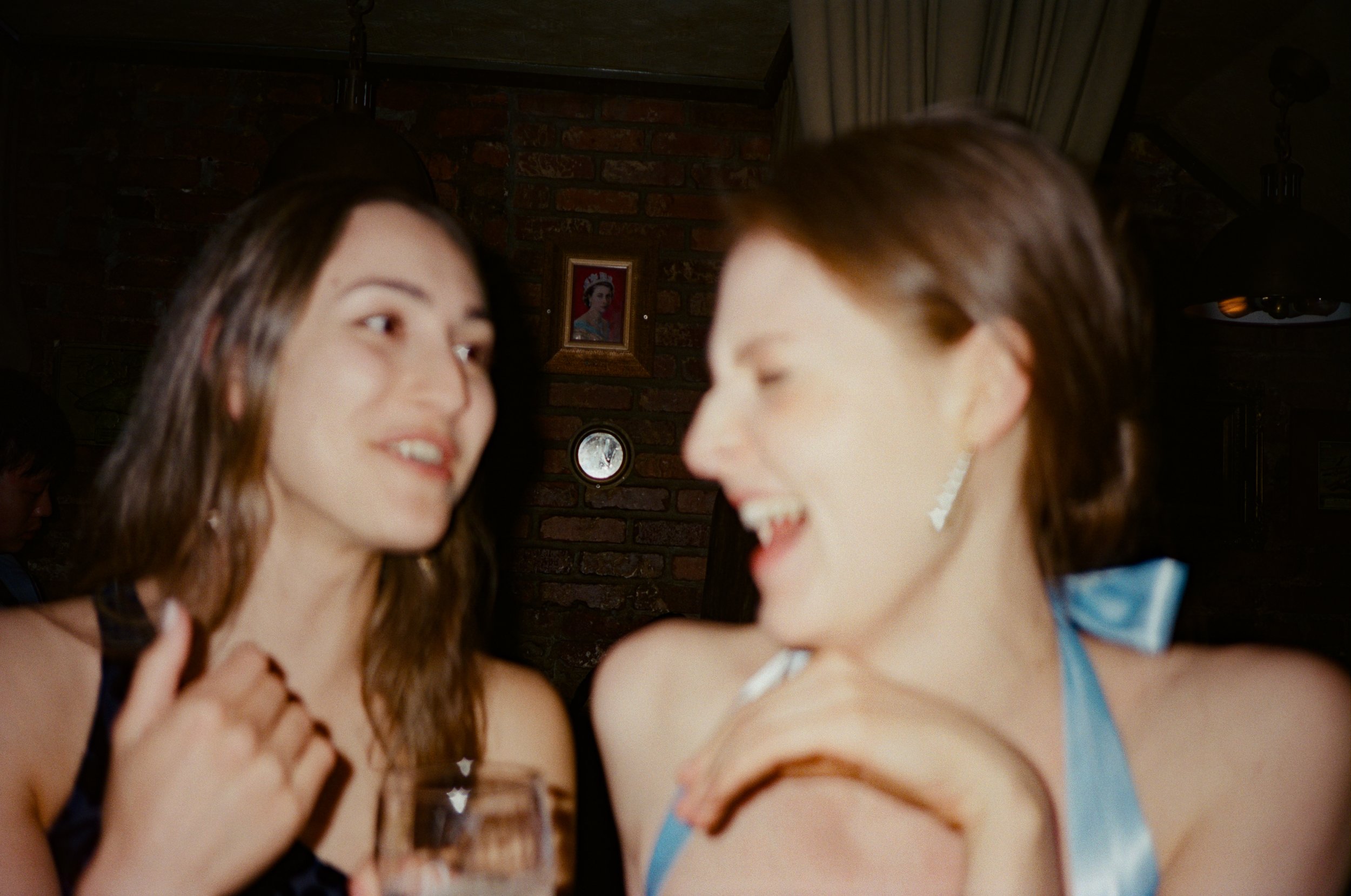 Two women laughing and talking closely in a dimly lit room with exposed brick wall, framed picture, and curtain in background.