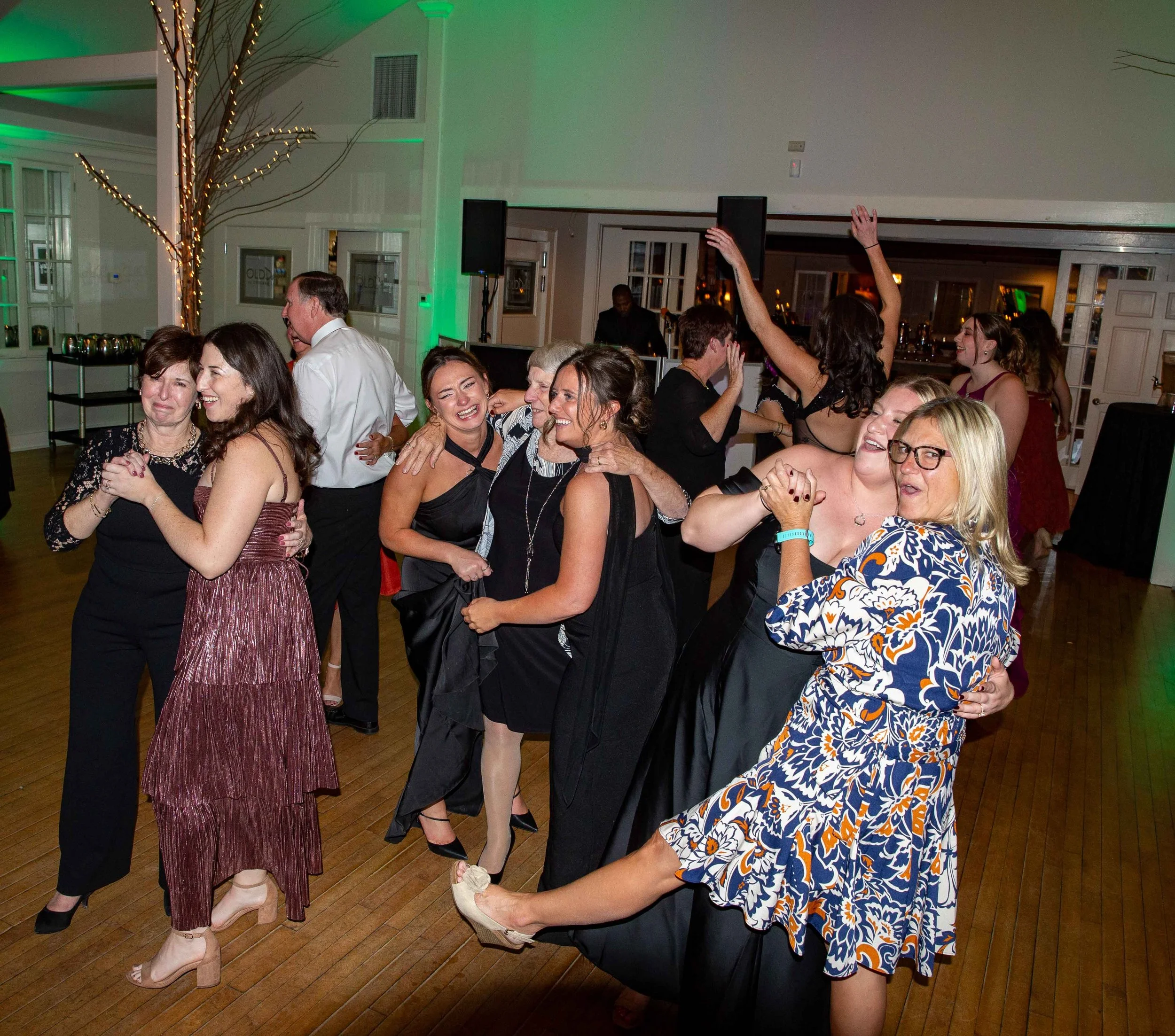 Group of people dancing and celebrating at a party or wedding reception in a decorated venue with string lights, green stage lighting, and a bar in the background.