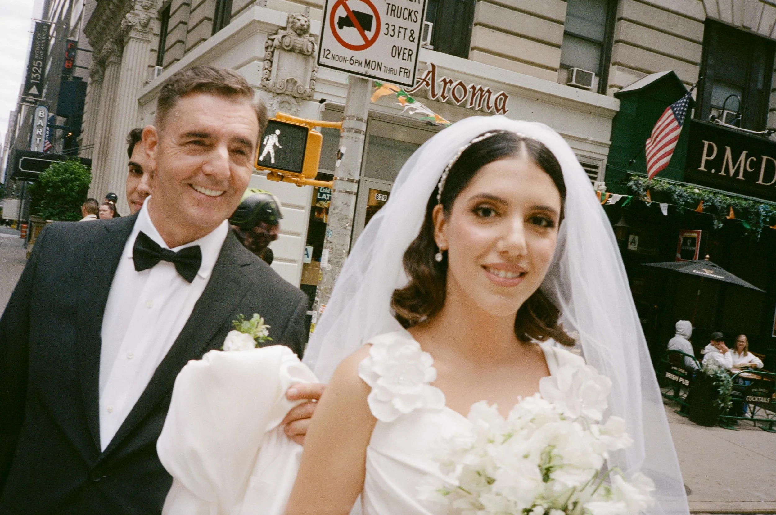 A bride in a white wedding dress with floral details on her shoulder, holding a bouquet of white flowers, and wearing a veil and pearl earrings. She stands next to a smiling man in a black tuxedo and bow tie on a city street, with buildings and peopl