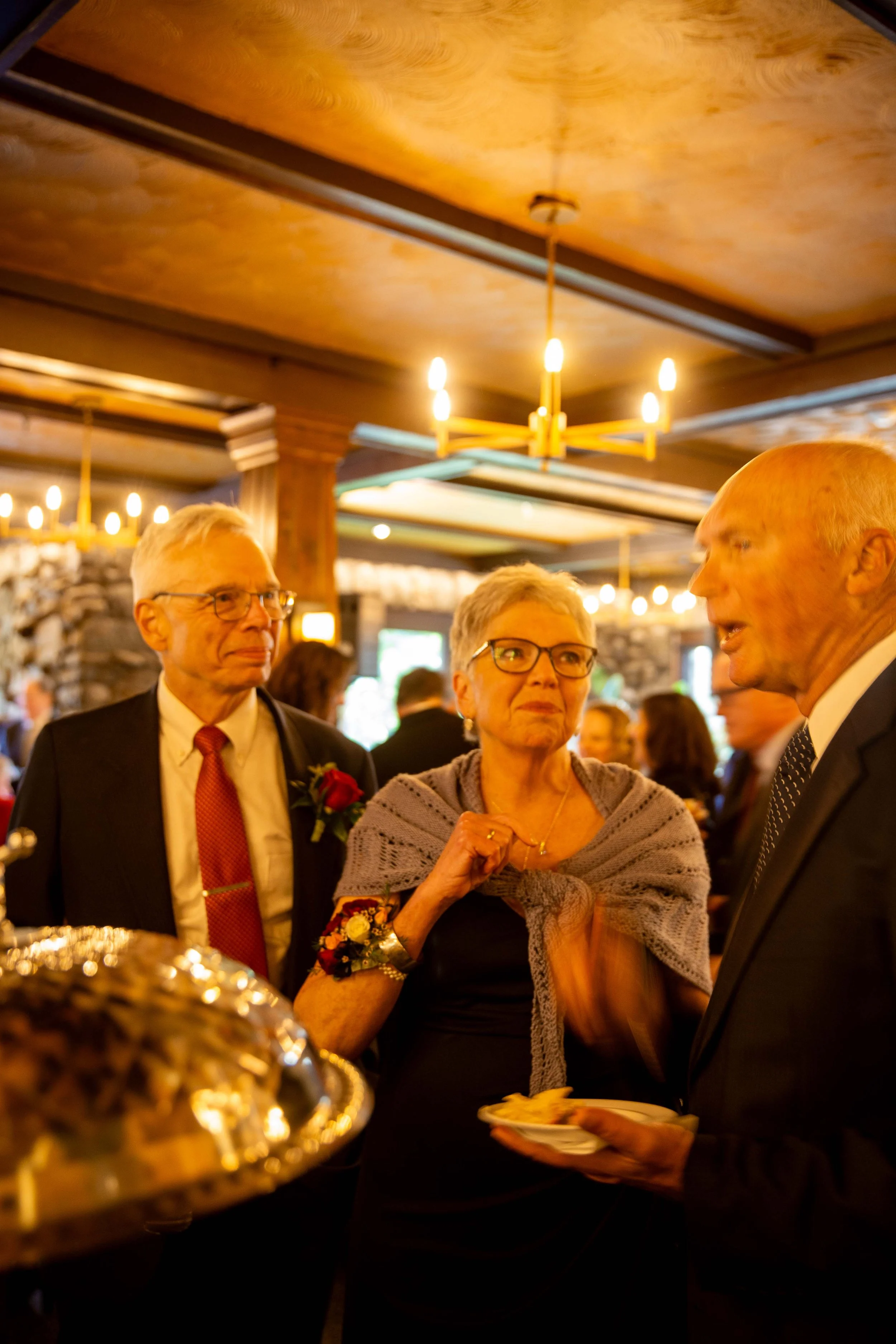 Three elderly adults in formal attire engaged in conversation at a social gathering in a warmly lit, rustic-style room with wooden ceiling beams and stone walls.
