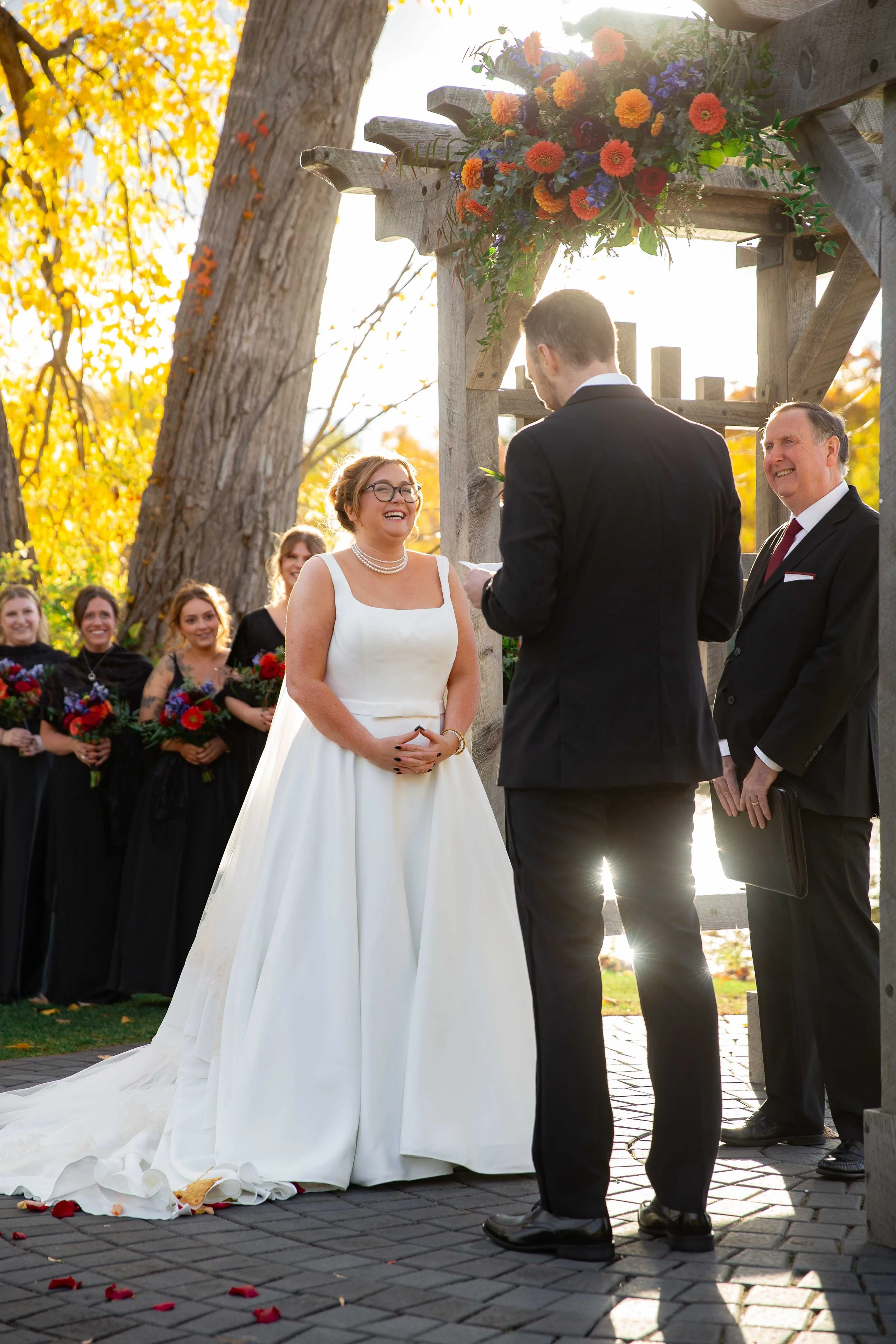 A wedding ceremony taking place outdoors under a wooden arch decorated with flowers, with a bride in a white dress and glasses looking at her groom, a man in a black suit, and an officiant. Bridesmaids in black dresses holding bouquets stand in the b