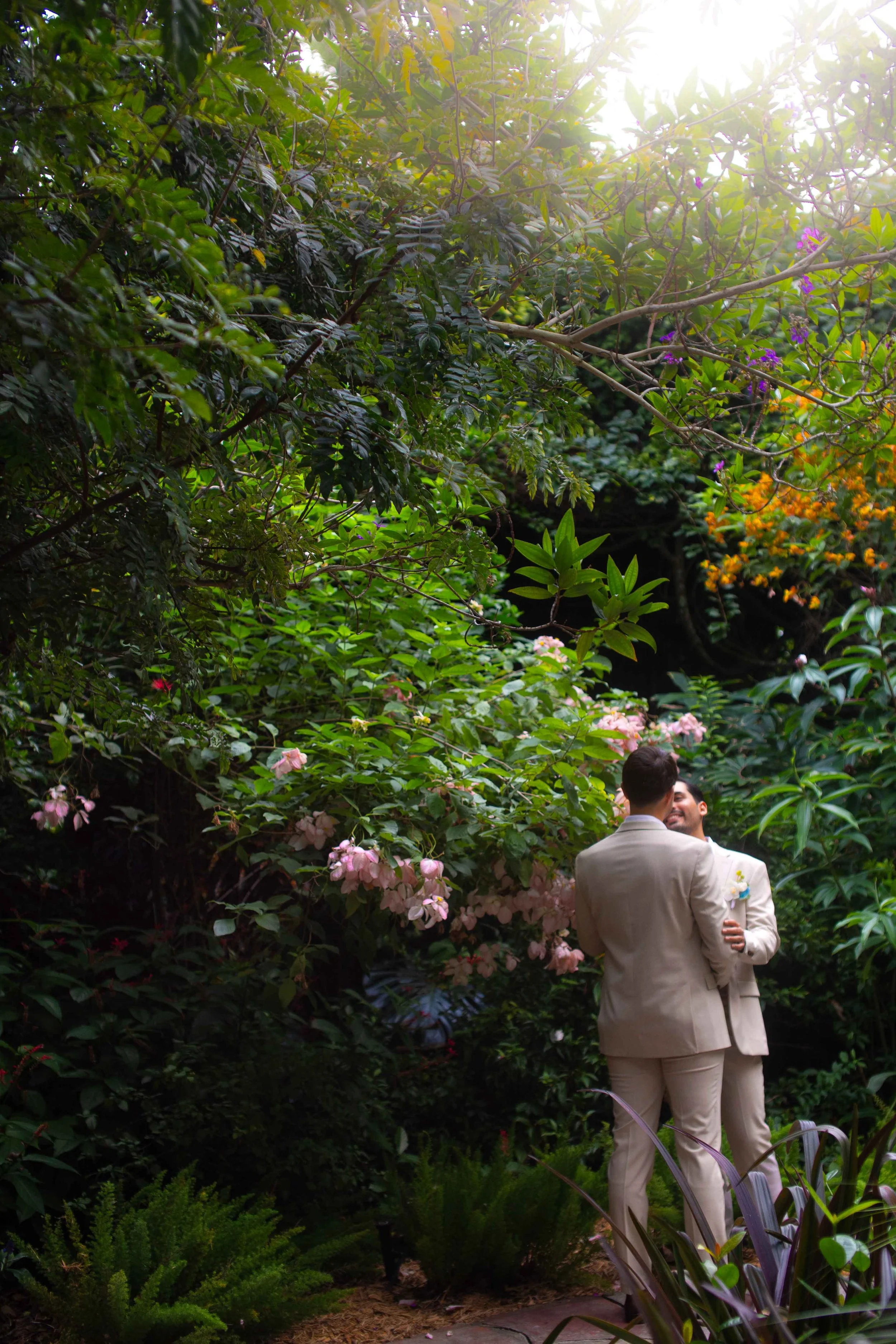 Two men in beige suits embracing and smiling in a lush garden with pink and purple flowers and greenery, with bright sunlight filtering through the trees.