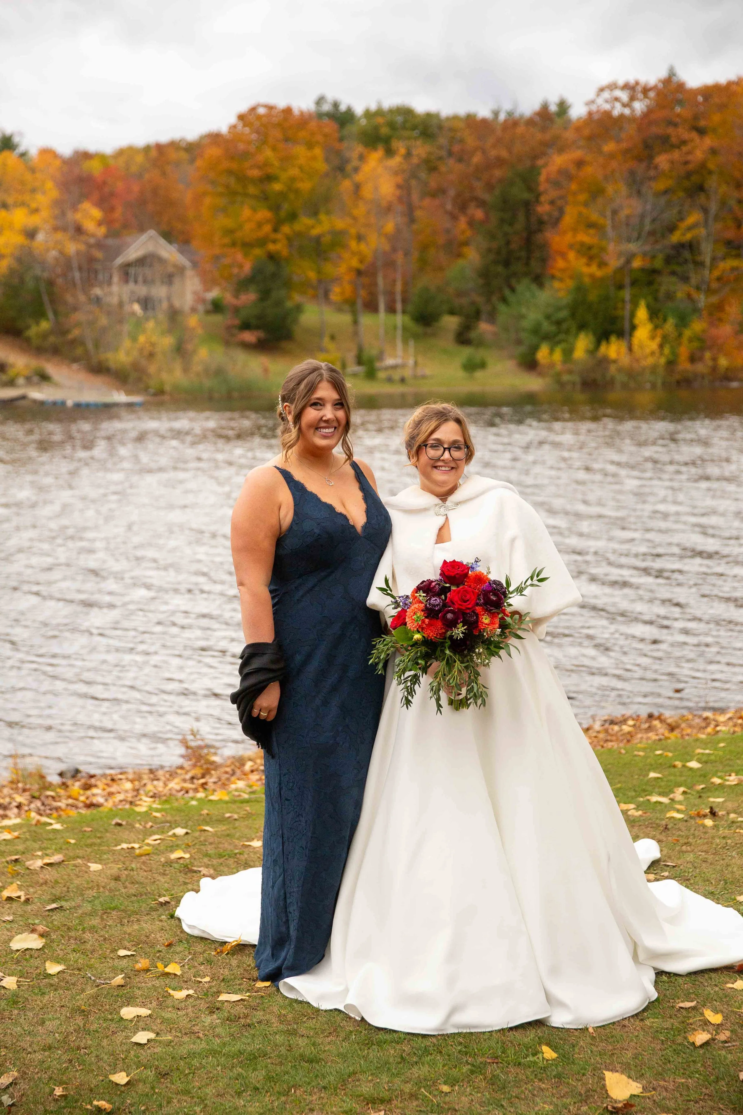Two women standing outdoors by a lake surrounded by fall foliage. One is wearing a navy blue dress, and the other is in a white wedding gown holding a bouquet of red, purple, and orange flowers.