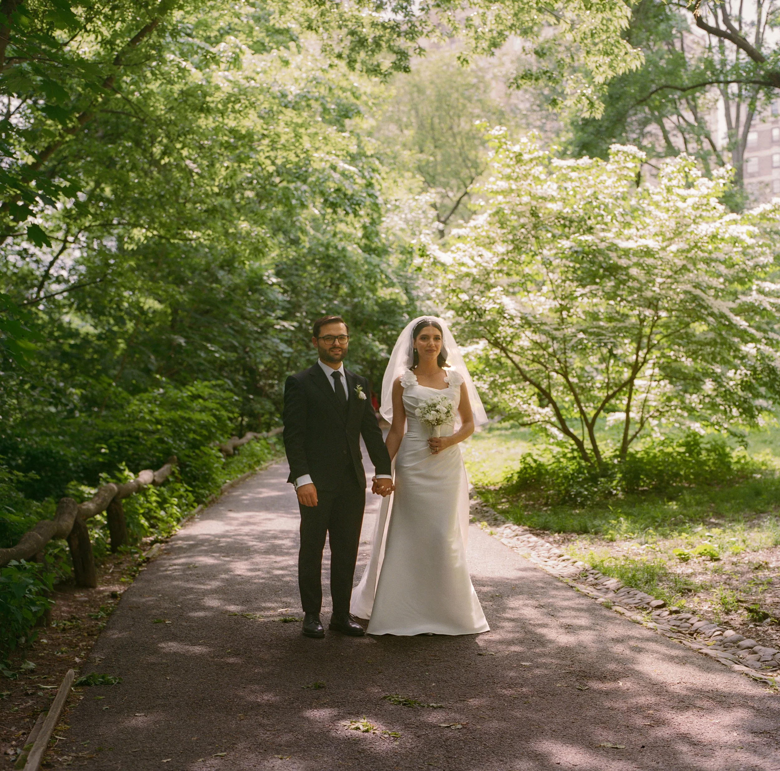 A bride and groom standing hand in hand on a tree-lined park path during daytime, with lush green foliage and white flowering trees in the background.