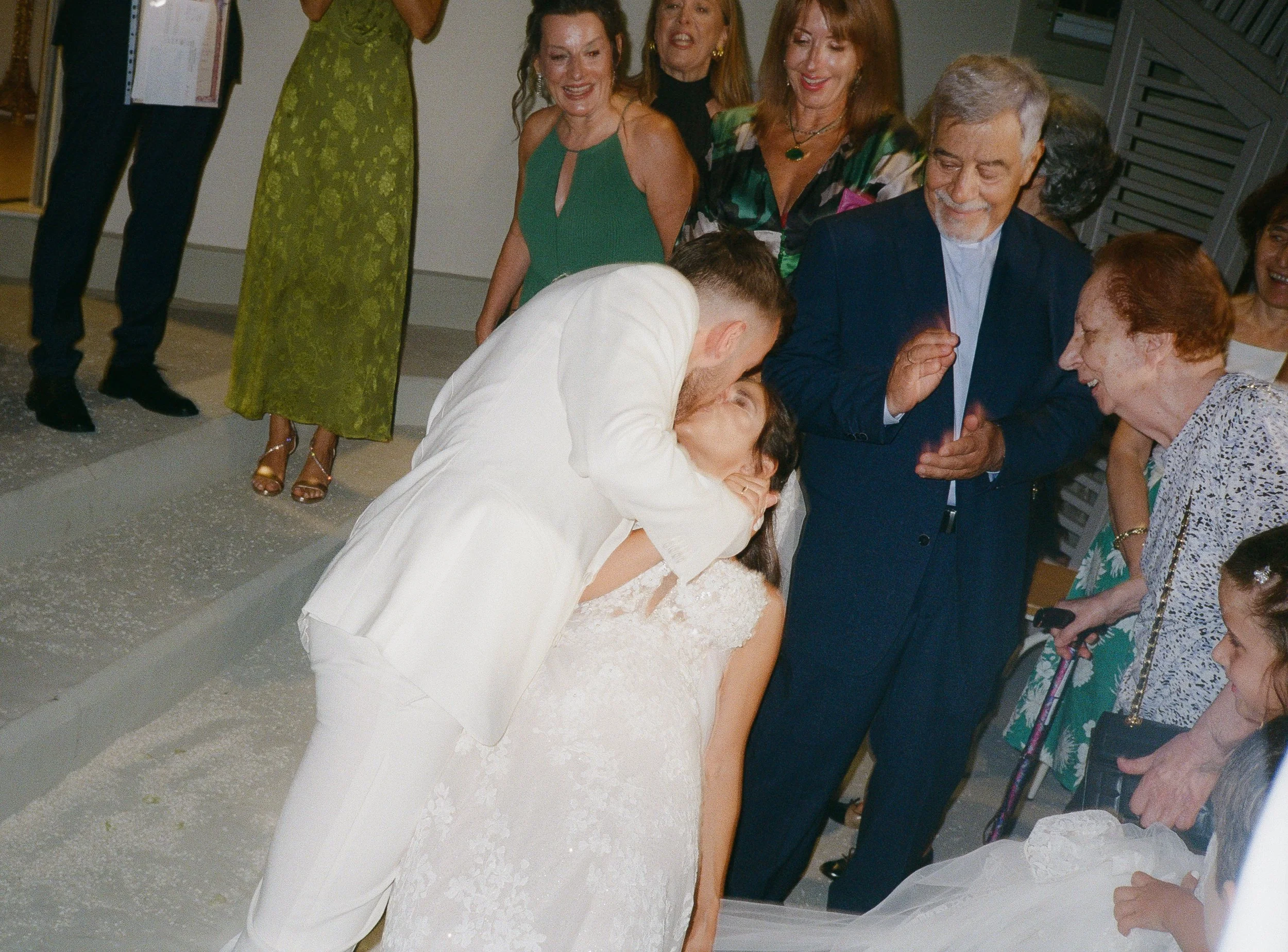 A wedding scene with a bride and groom kissing, surrounded by smiling family and friends.