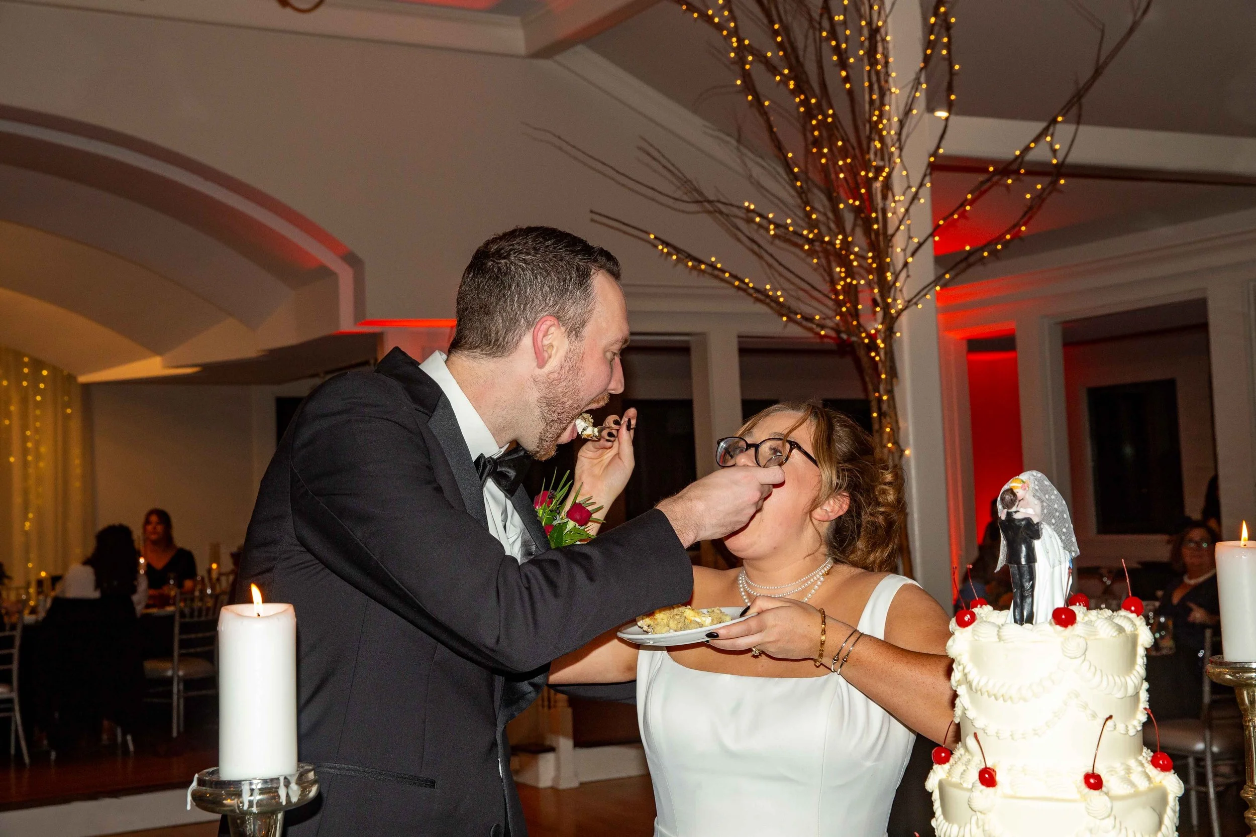 A bride and groom share a cake at their wedding reception. The groom wears a tuxedo and feeds the bride a piece of cake. The bride is wearing glasses and a white wedding dress, with her hand holding a plate. There is a wedding cake decorated with che
