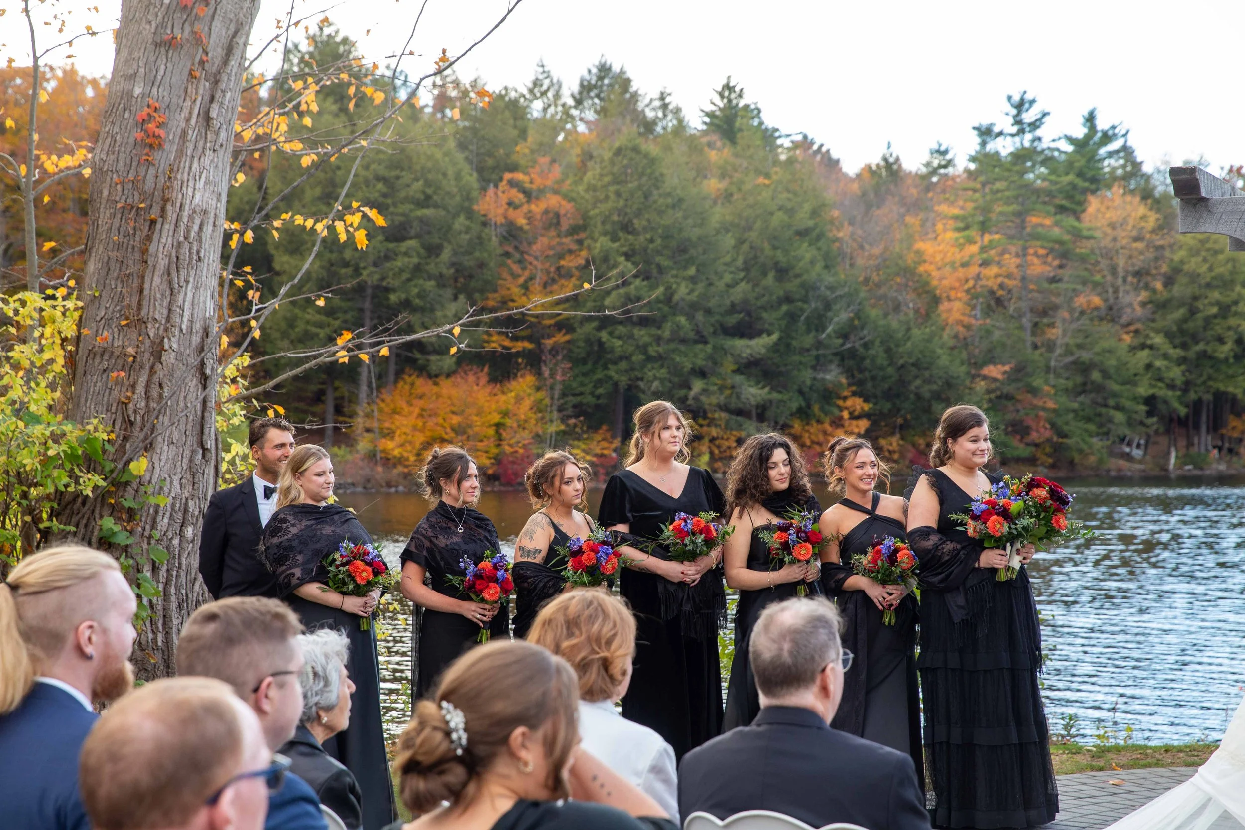 A wedding ceremony taking place outdoors by a lake in autumn, with bridesmaids and a groomsman standing in a line, holding bouquets of flowers, facing the water, with guests seated in front. The background shows colorful fall foliage and trees.