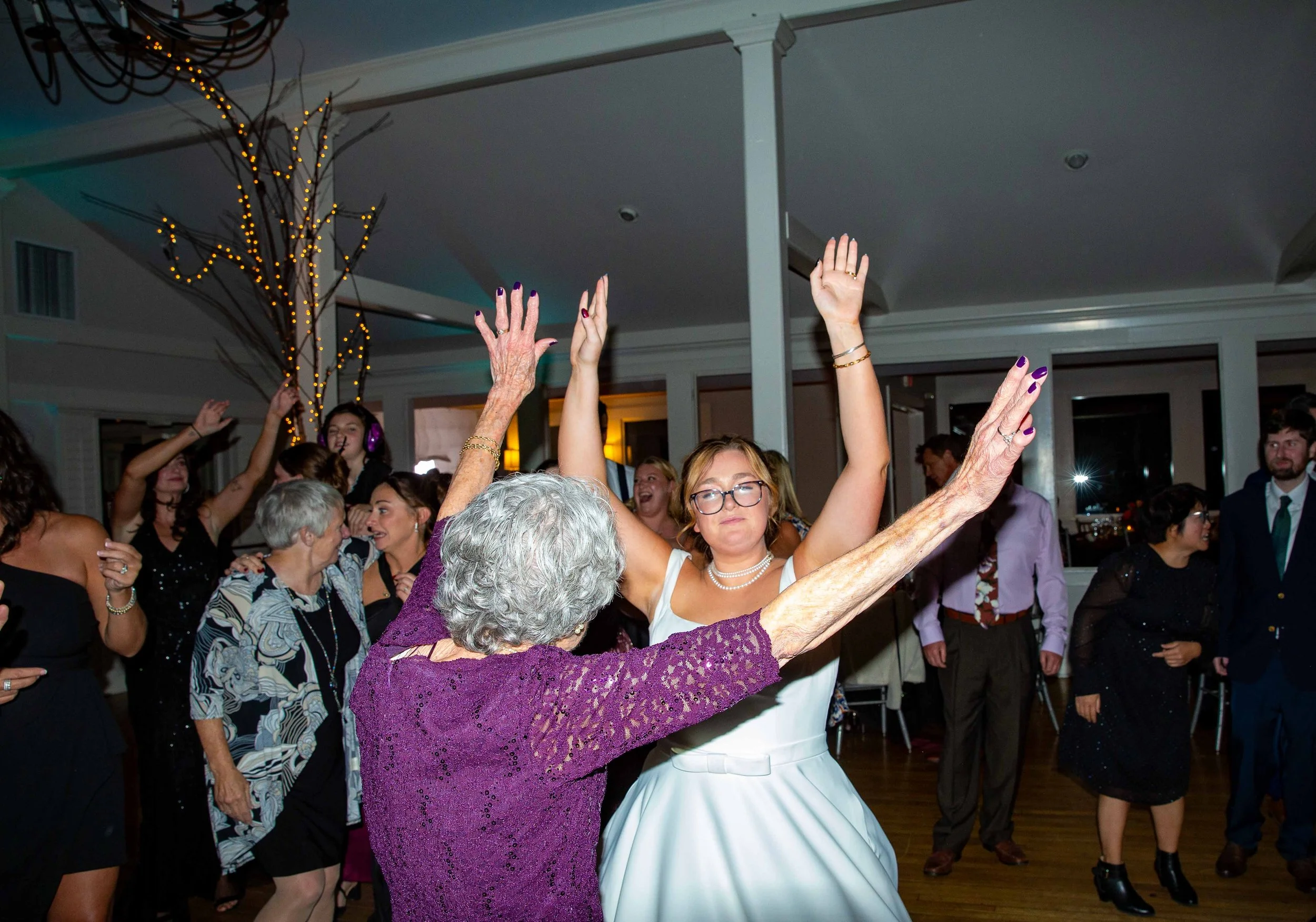 An elderly woman and a young woman are dancing together at a celebration or wedding reception, with other guests dancing and celebrating around them in a decorated indoor venue.