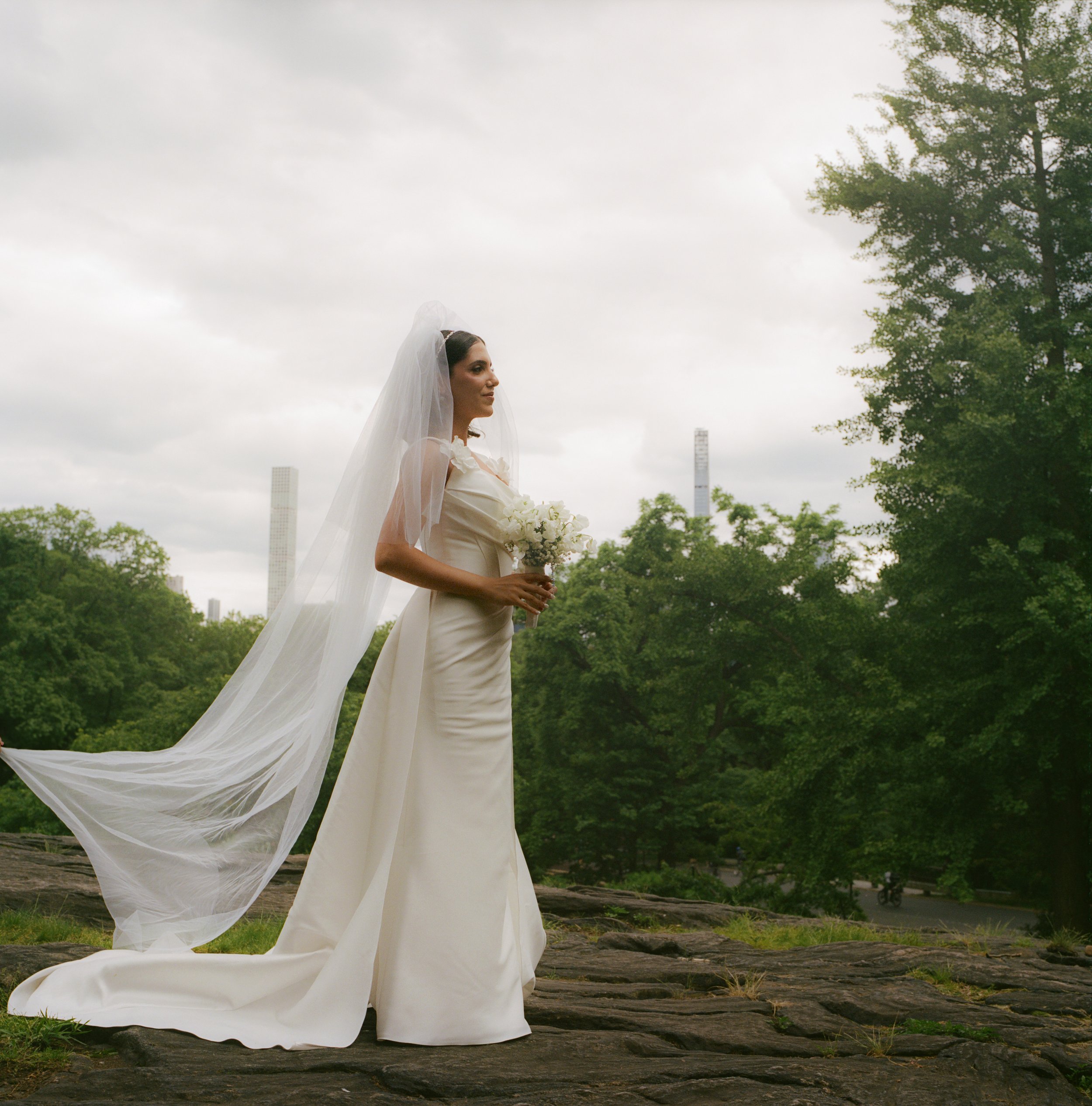 A bride in a white wedding dress with a long train and veil standing outdoors on a rocky surface, holding a bouquet of white flowers, with trees and tall buildings in the background on a cloudy day.