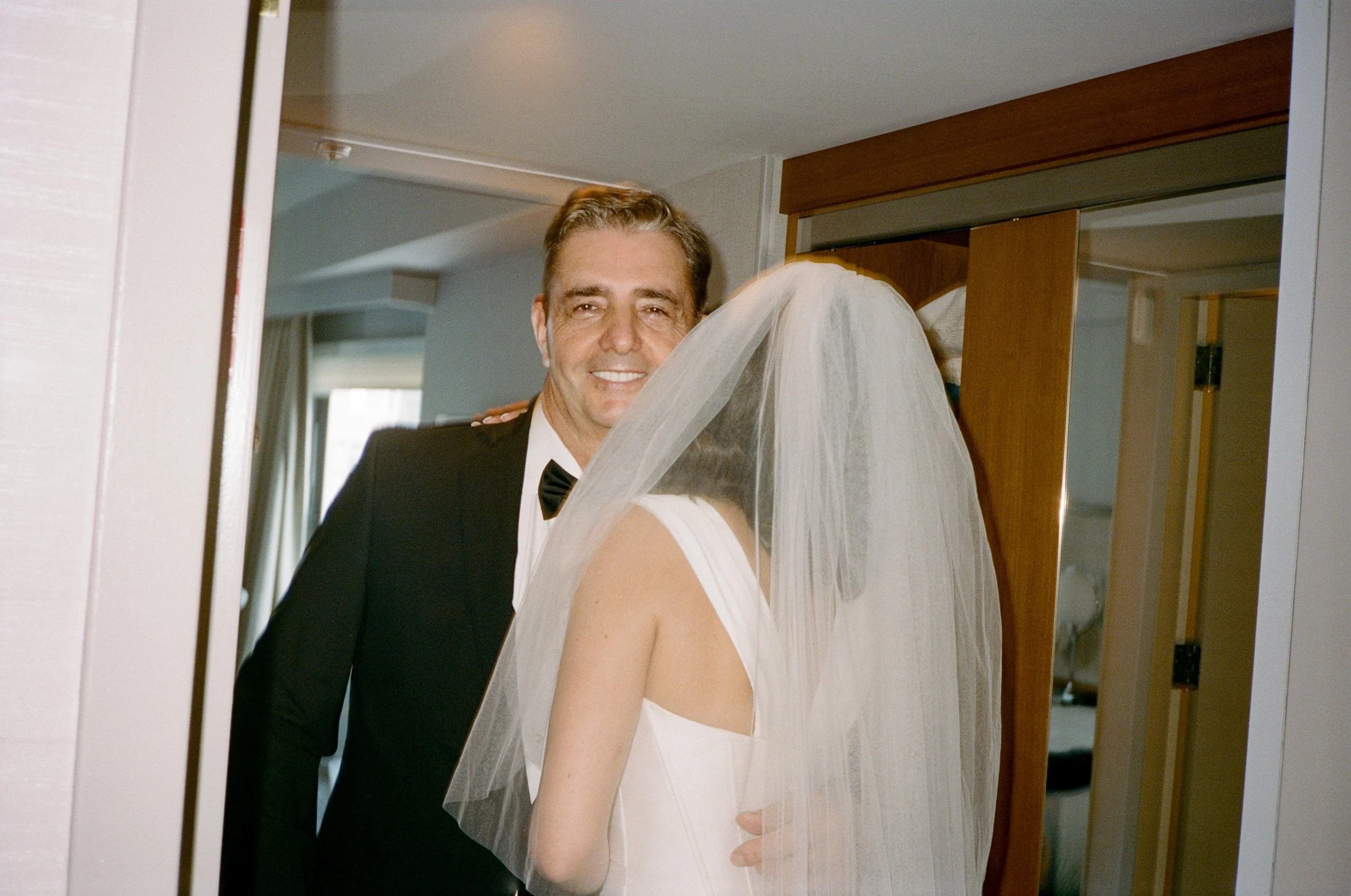 A man in a tuxedo smiling and greeting a bride with a veil in an indoor setting, likely a hotel room.