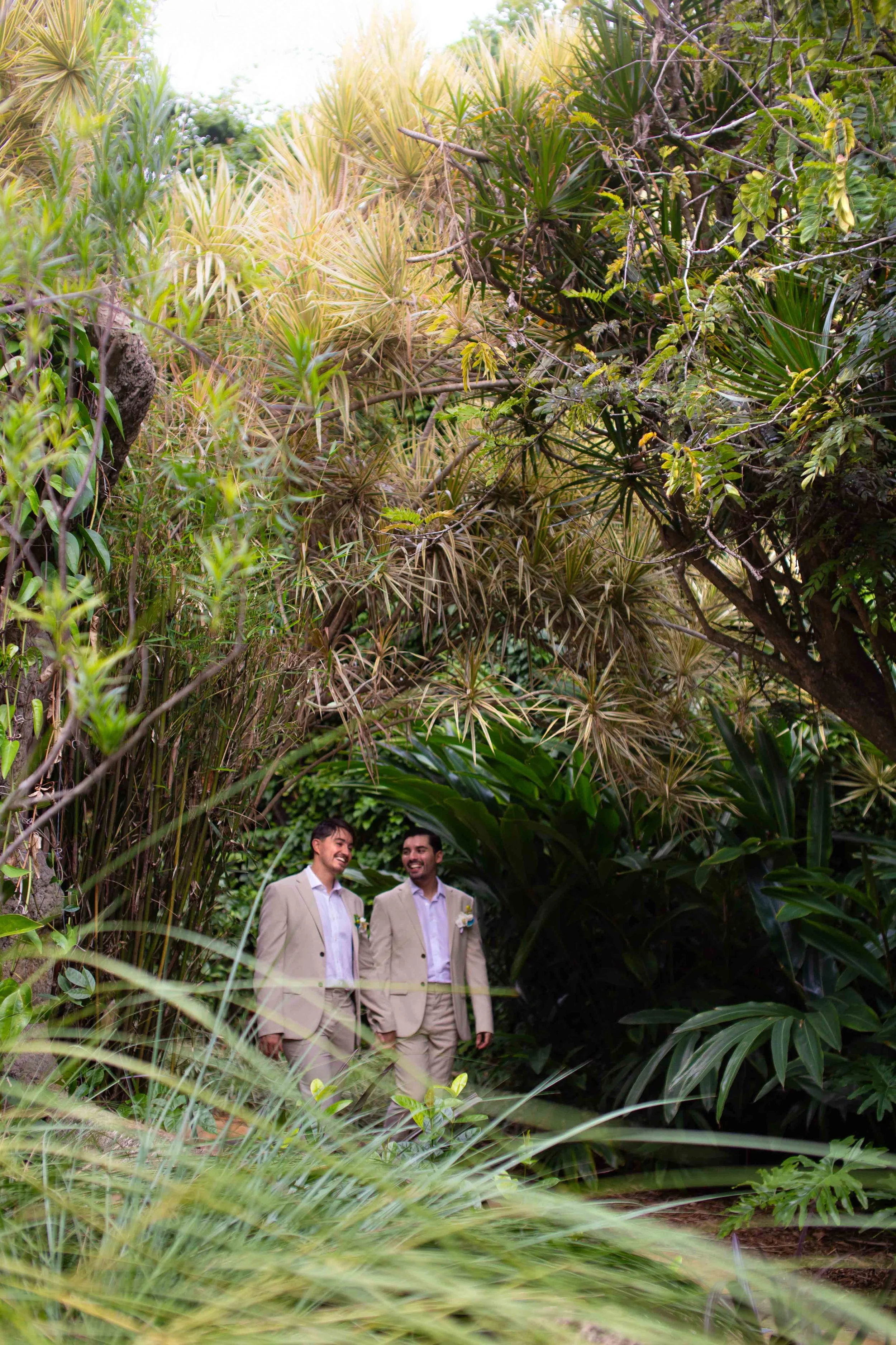 Two men dressed in beige suits standing amidst lush green tropical plants, smiling and talking.