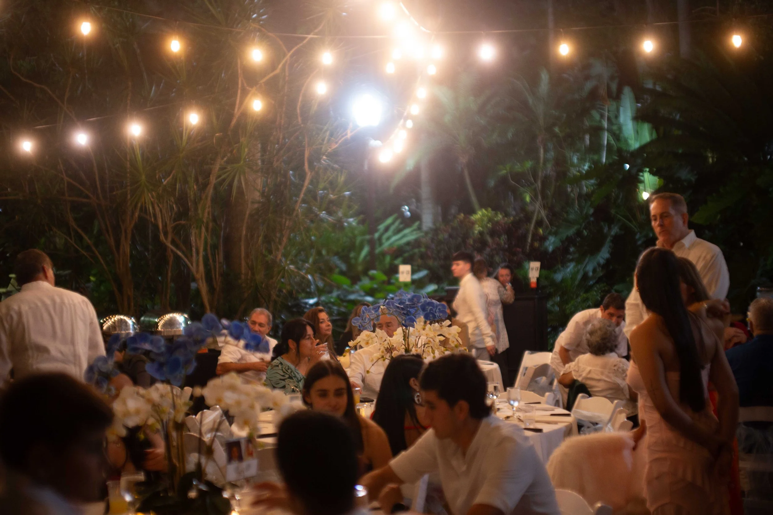 A group of people gathered at a nighttime outdoor celebration or party, illuminated by string lights overhead, with lush greenery in the background. Guests are seated at tables decorated with white floral centerpieces, and some people are standing an