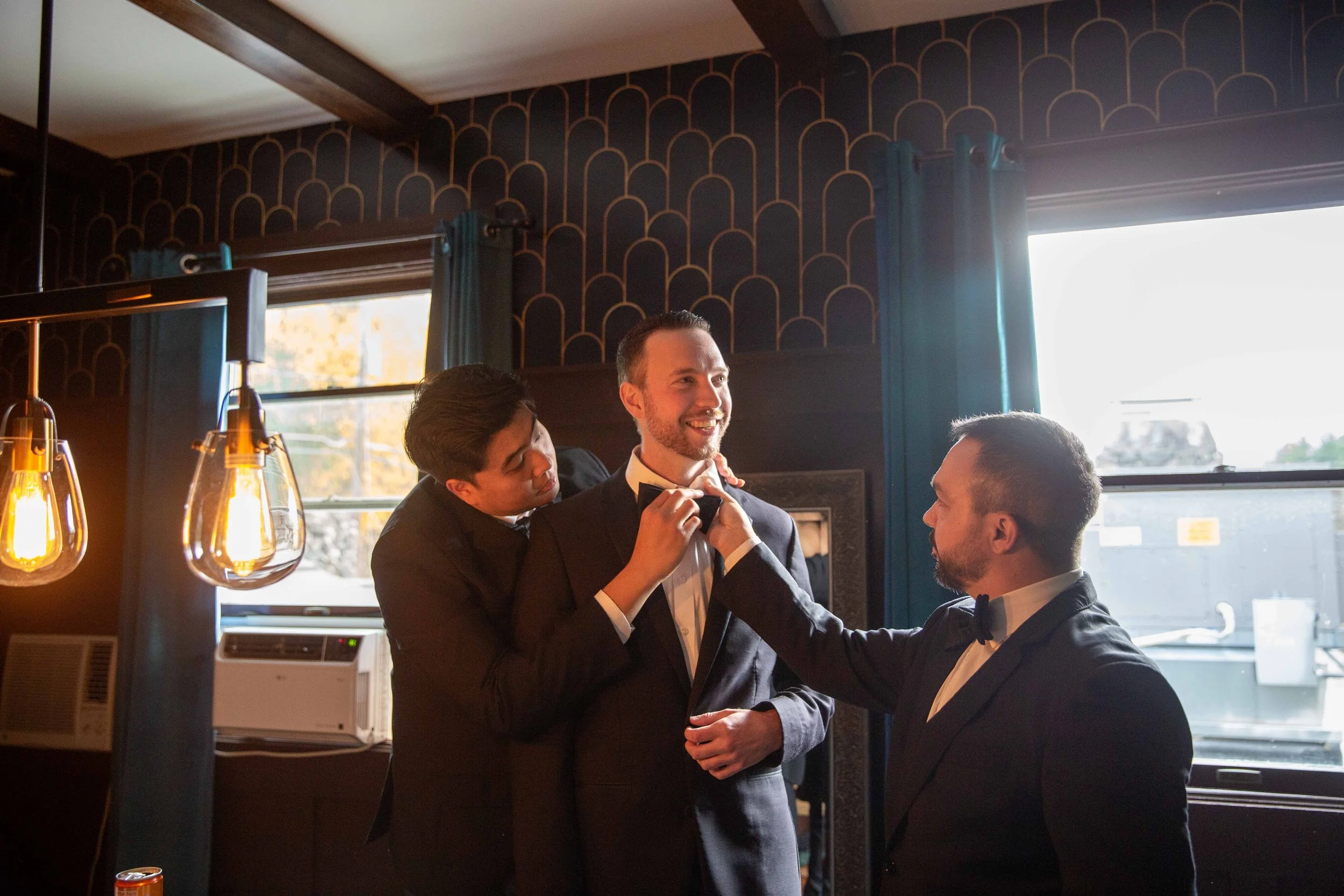 Three men in formal suits, two men adjusting the bow tie of the third man in the middle, inside a room with dark blue walls, pendant lights, and large windows