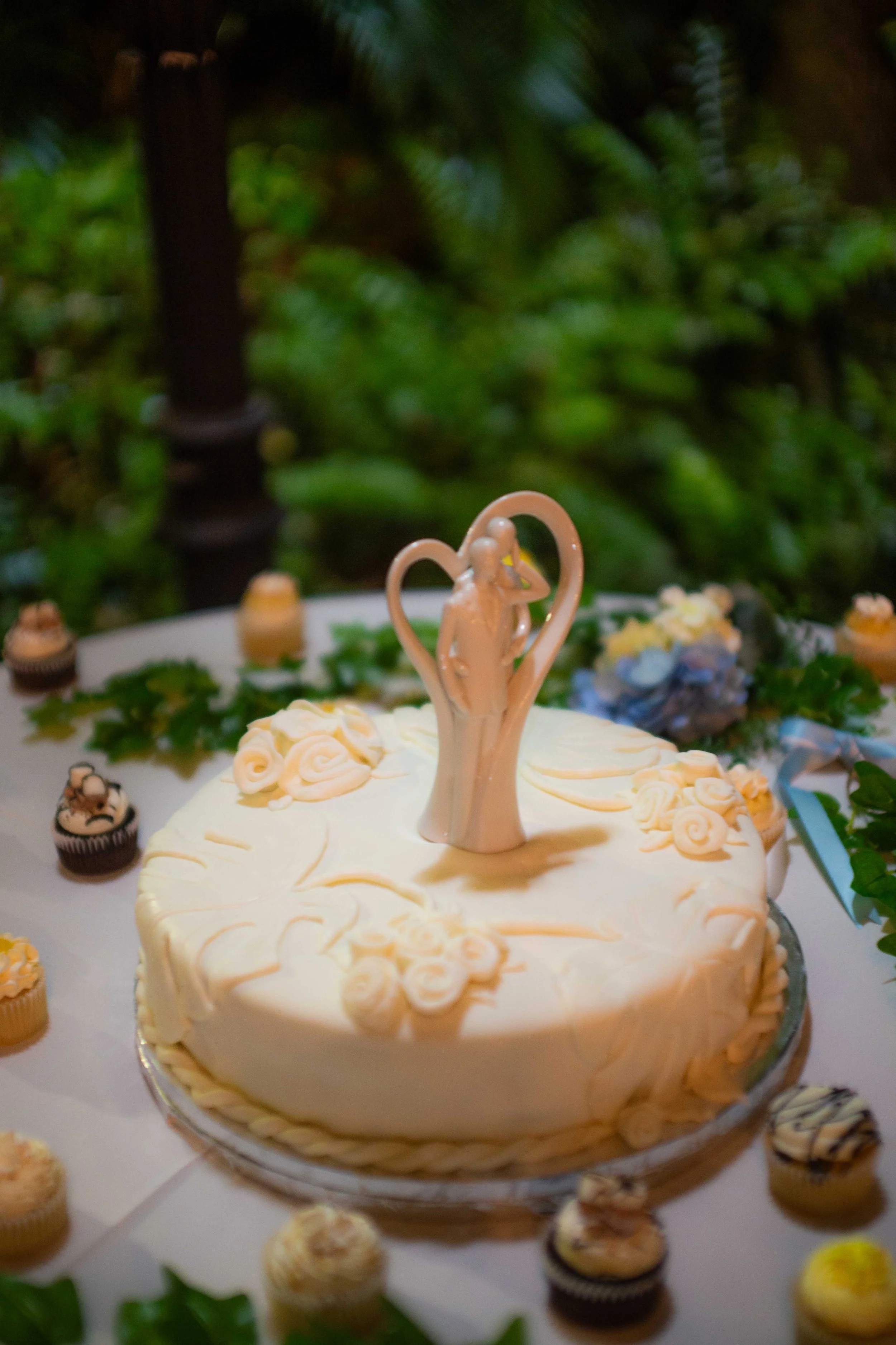 A white wedding cake decorated with white roses and a bride and groom figurine topper, surrounded by cupcakes, on a table with greenery.