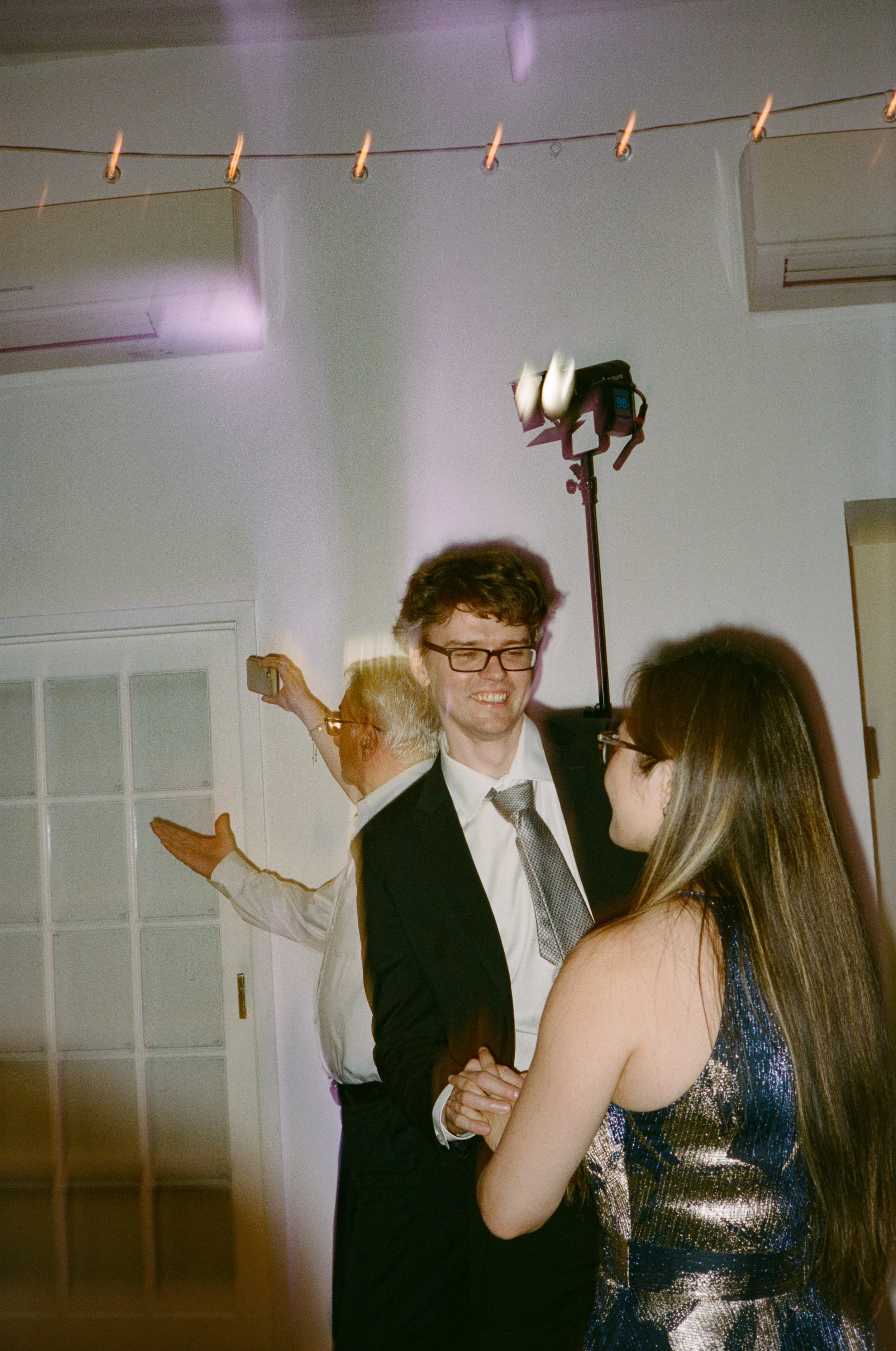 People dancing at a party, with a man in a suit and glasses smiling and a woman with long hair in a sparkly dress, in a room with white walls, air conditioning units, and party lights.