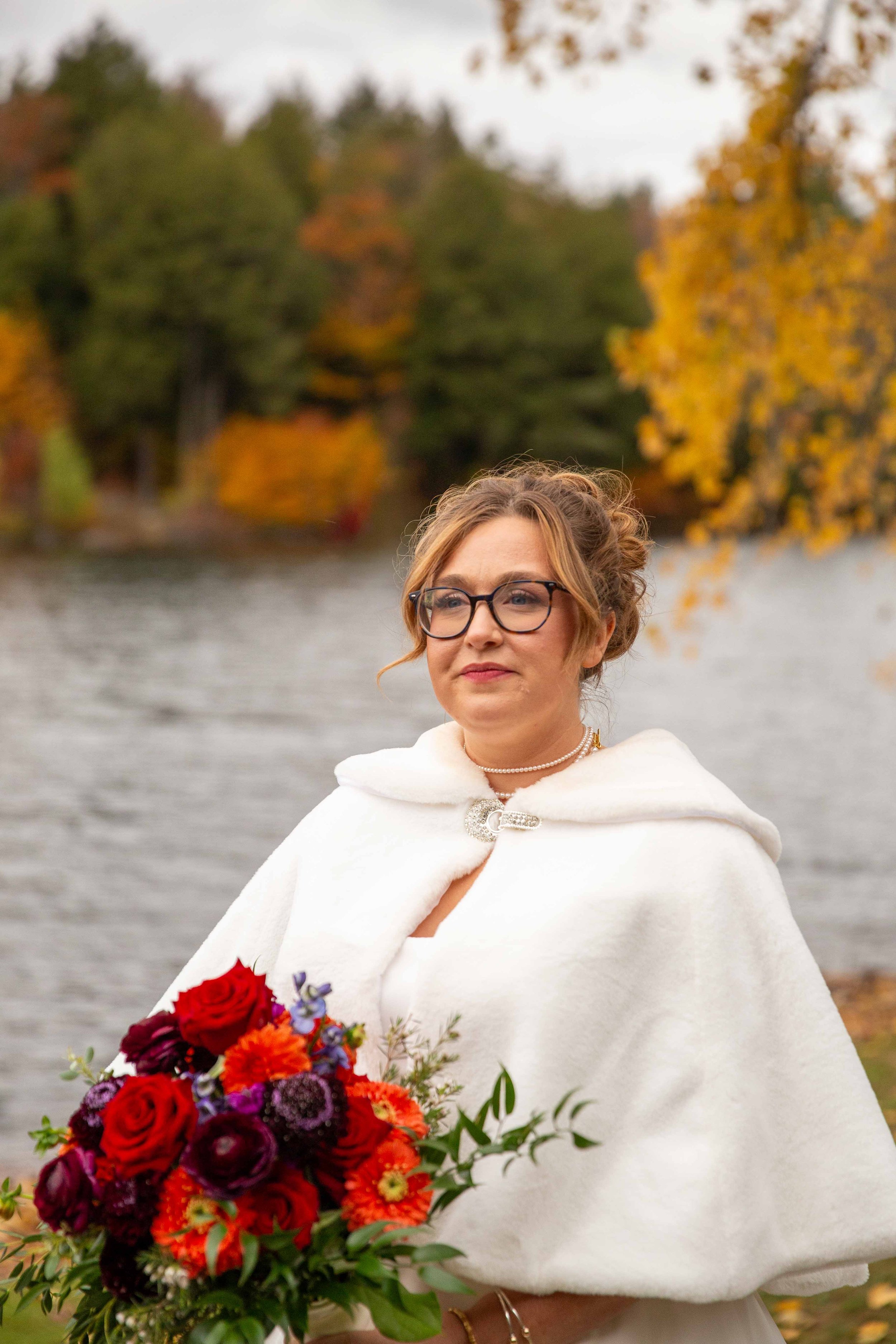 A woman wearing glasses and a white cape holding a colorful bouquet of flowers near a lake with autumn trees in the background.