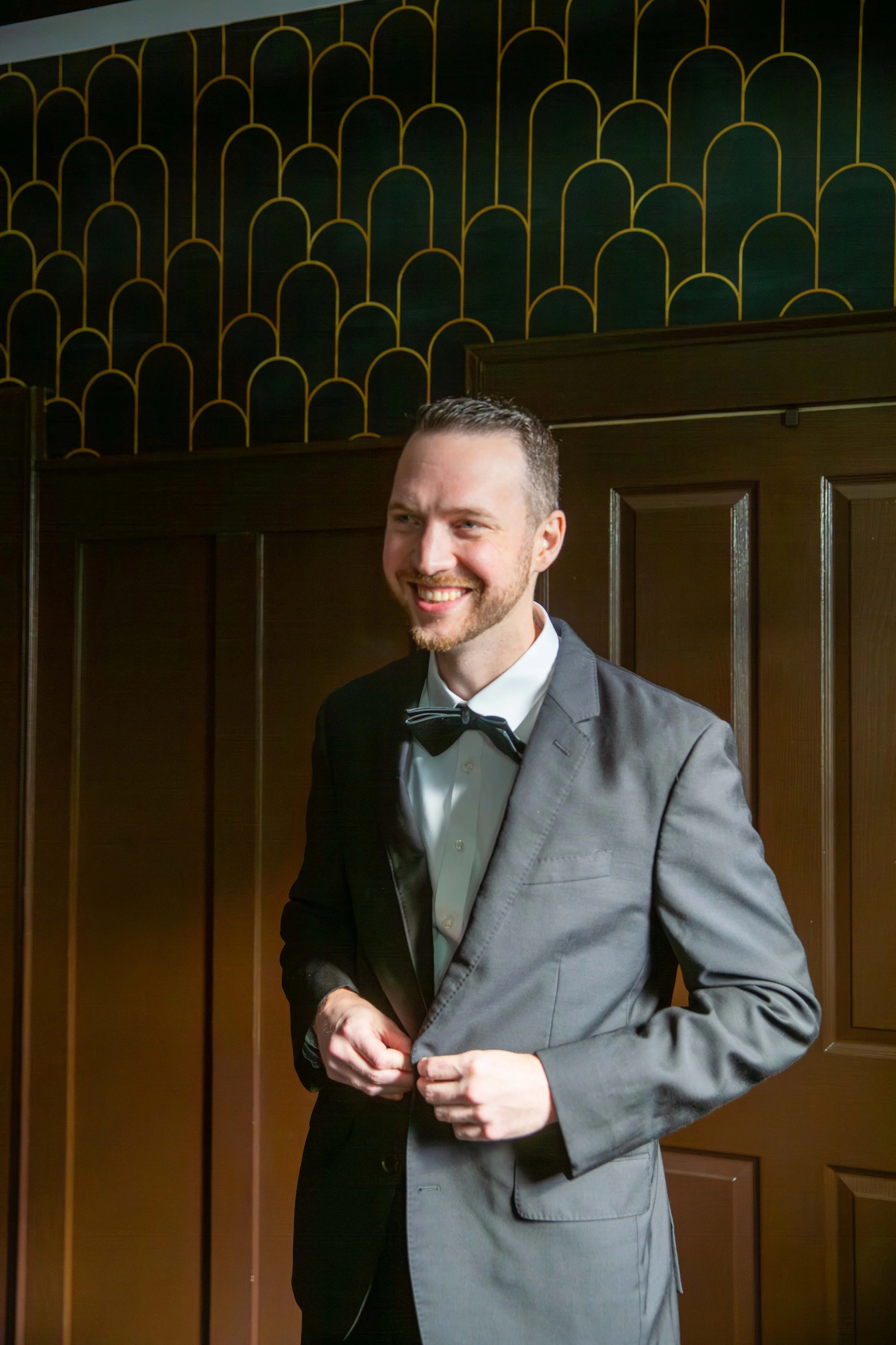 A man in a gray tuxedo with a black bow tie, smiling, adjusting his jacket, standing in front of dark brown wooden doors and a green and gold decorative wall.