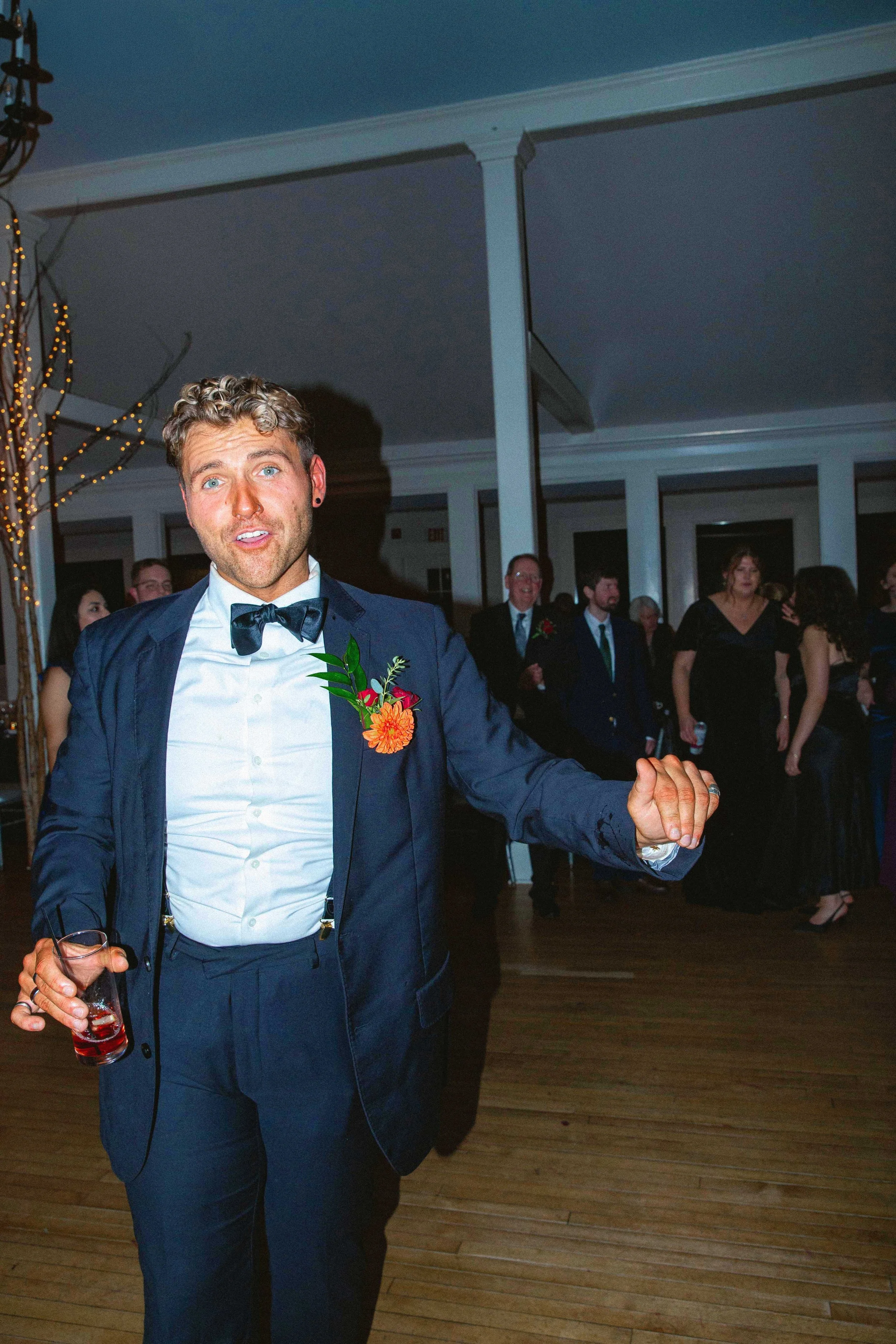 A man in a tuxedo dances at a party or wedding reception, holding a drink in his left hand, with other guests in formal attire in the background.