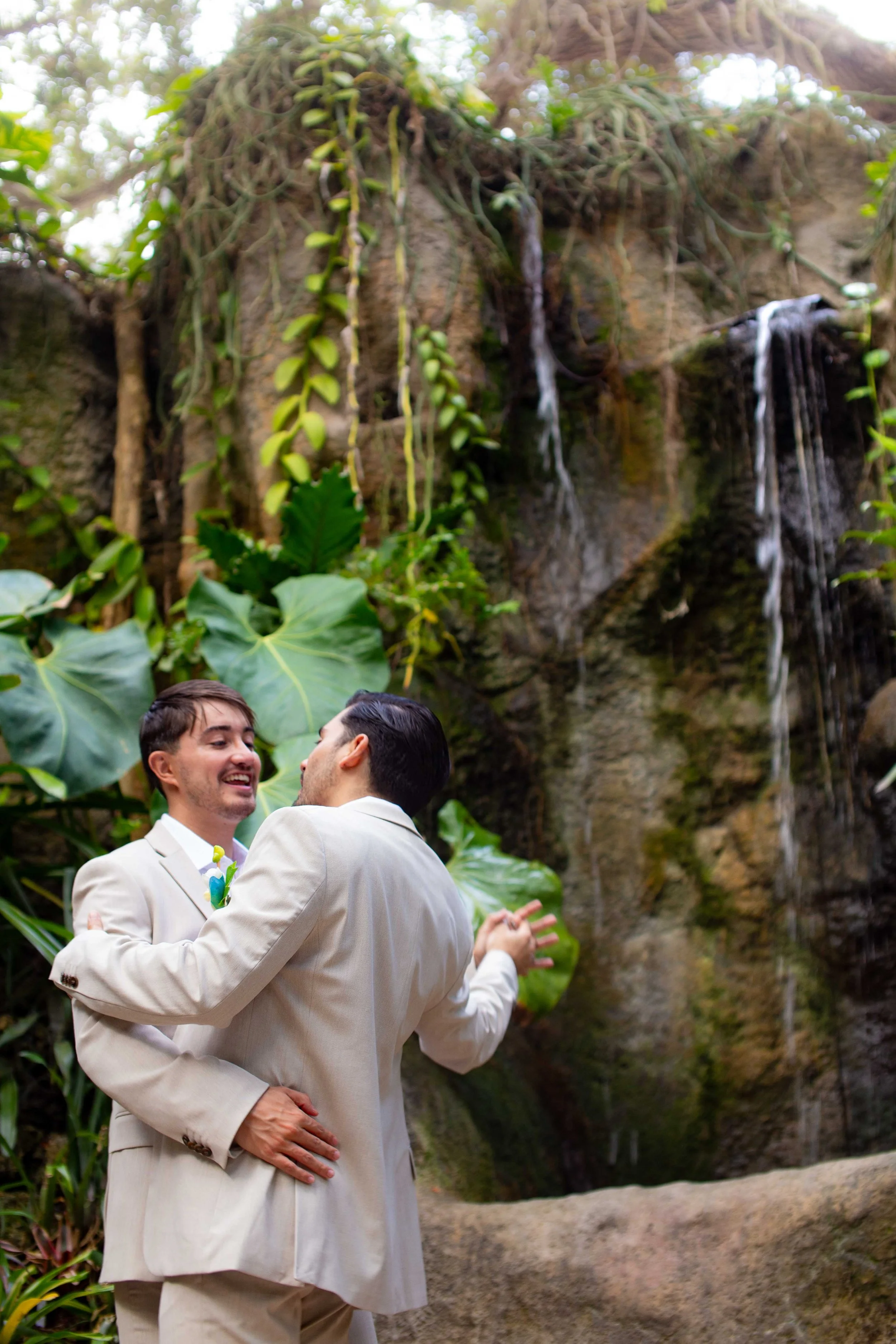 Two men in cream suits dancing and smiling in front of a waterfall in a lush, tropical setting.