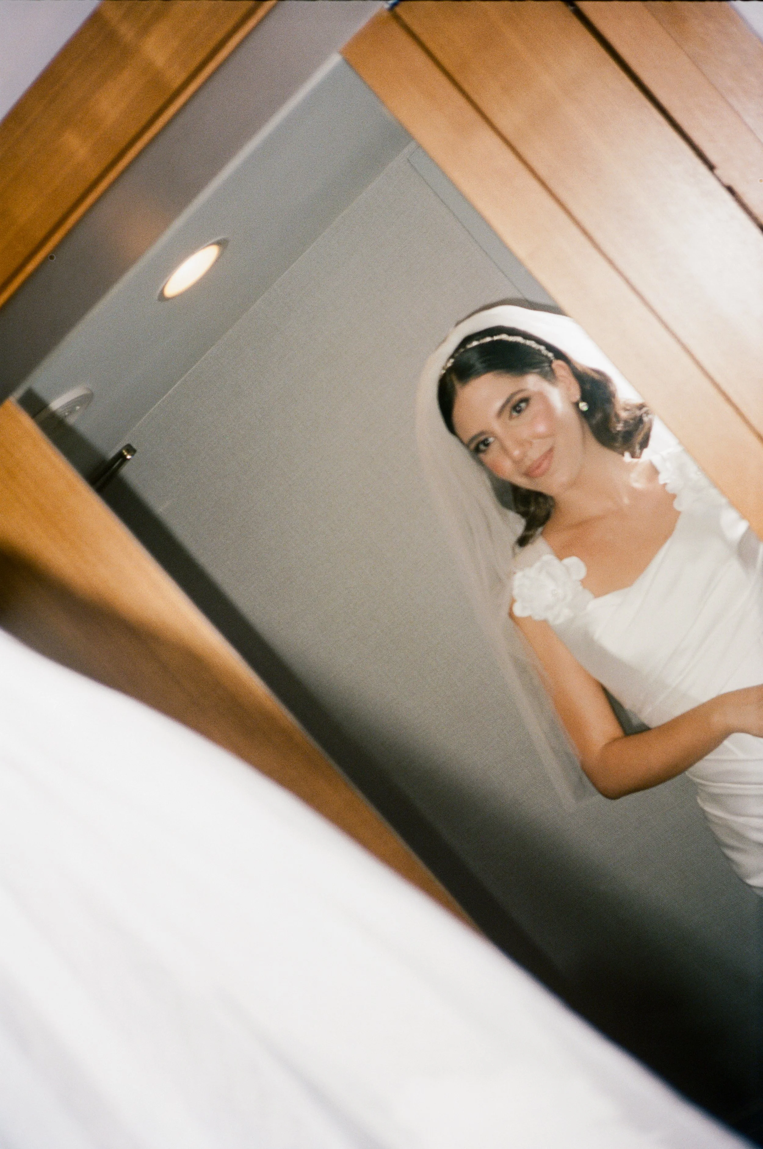 A woman in a white dress with floral details on the shoulders is looking into a mirror, with her reflection visible. She is wearing earrings and a headband with a veil, suggesting she is a bride.
